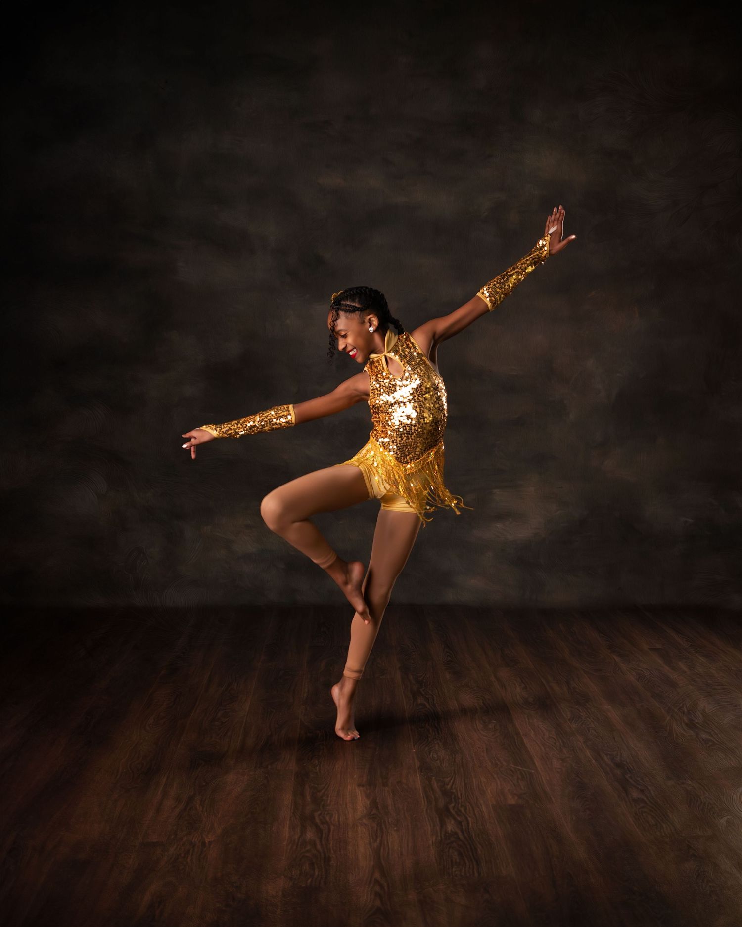 A dancer in a gold sequined fringe costume poses gracefully on one leg in a professional studio setting.