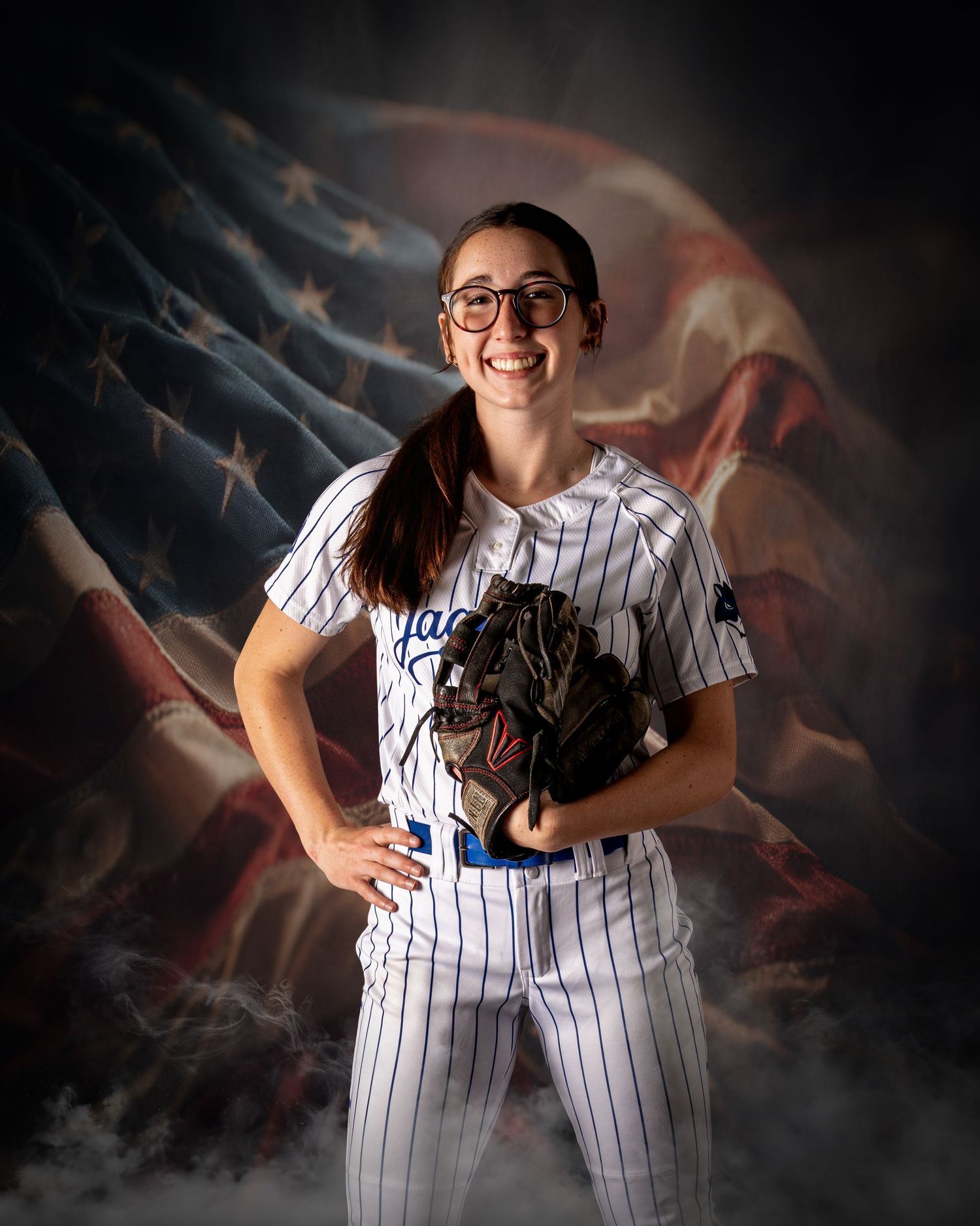 Smiling female softball player in pinstripe uniform holding glove, posing before American flag backdrop.
