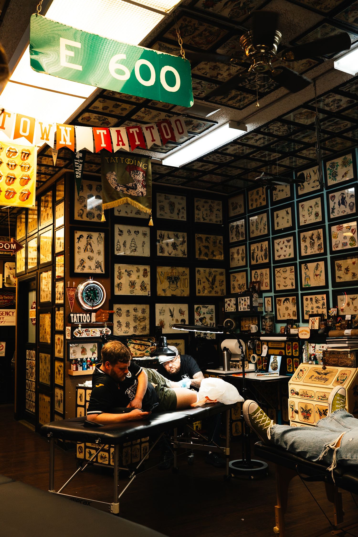 Interior of a tattoo shop with walls covered in flash art designs and an E 600 street sign.