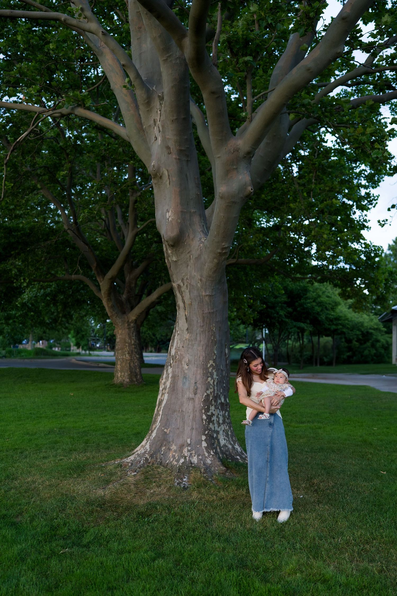 Person holding baby stands beneath large tree at dusk in park setting.