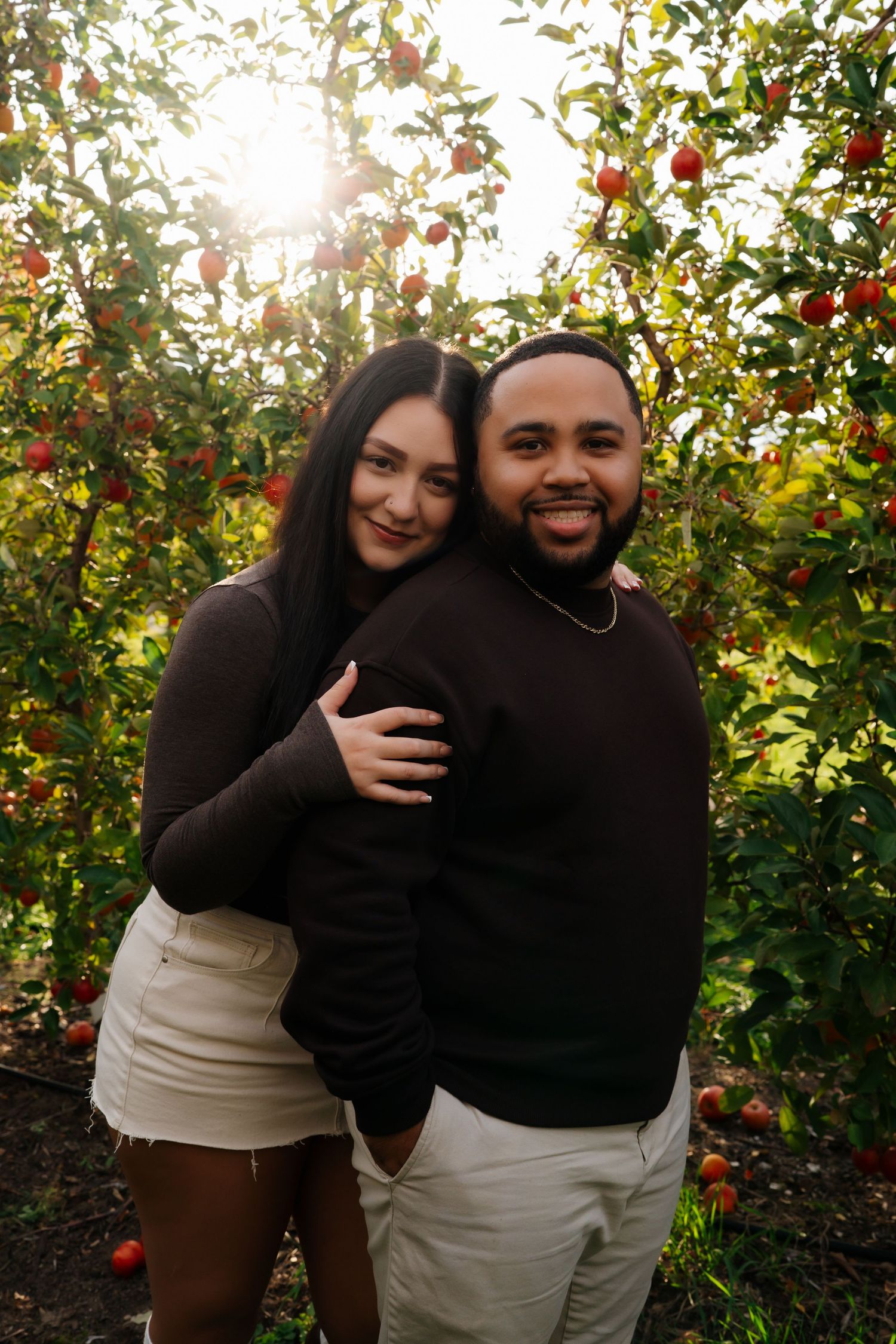 Couple embracing in apple orchard during golden hour with sun flare and red apples.