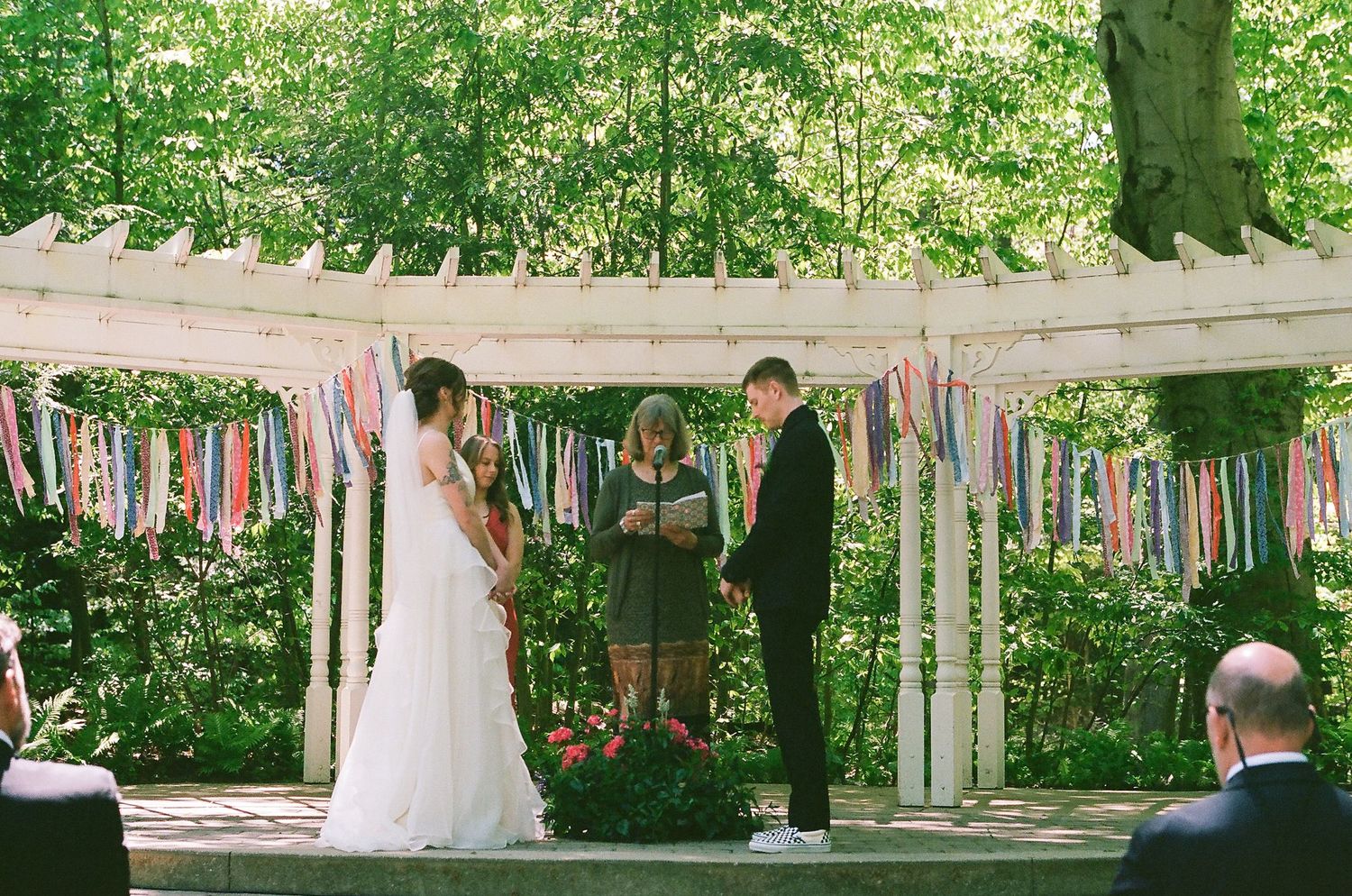 An outdoor wedding ceremony takes place under a rustic wooden arch decorated with colorful fabric streamers in a garden setting.