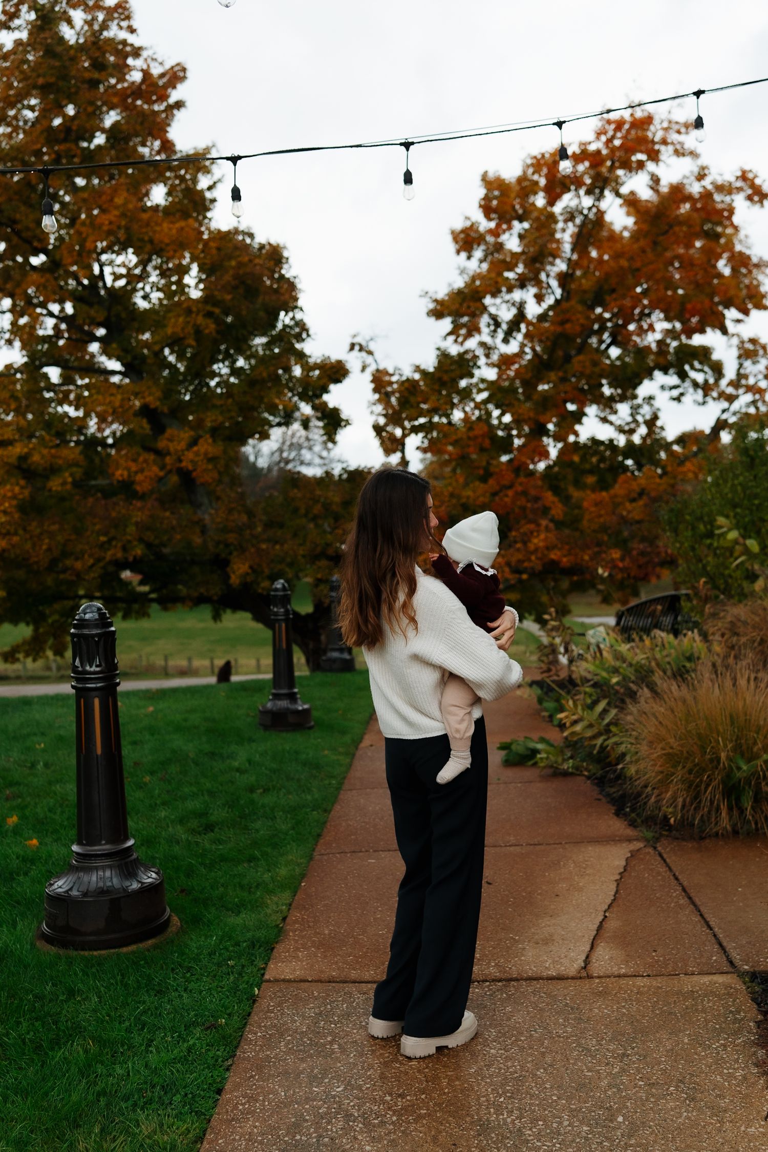 Person in white sweater holds baby while standing on pathway with string lights overhead.