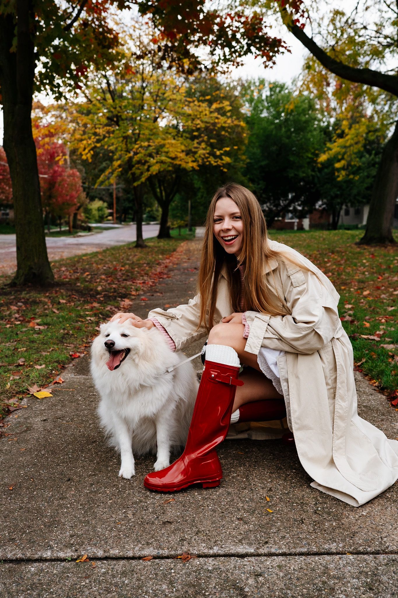 Happy person in trench coat kneels beside smiling white fluffy dog on autumn sidewalk.