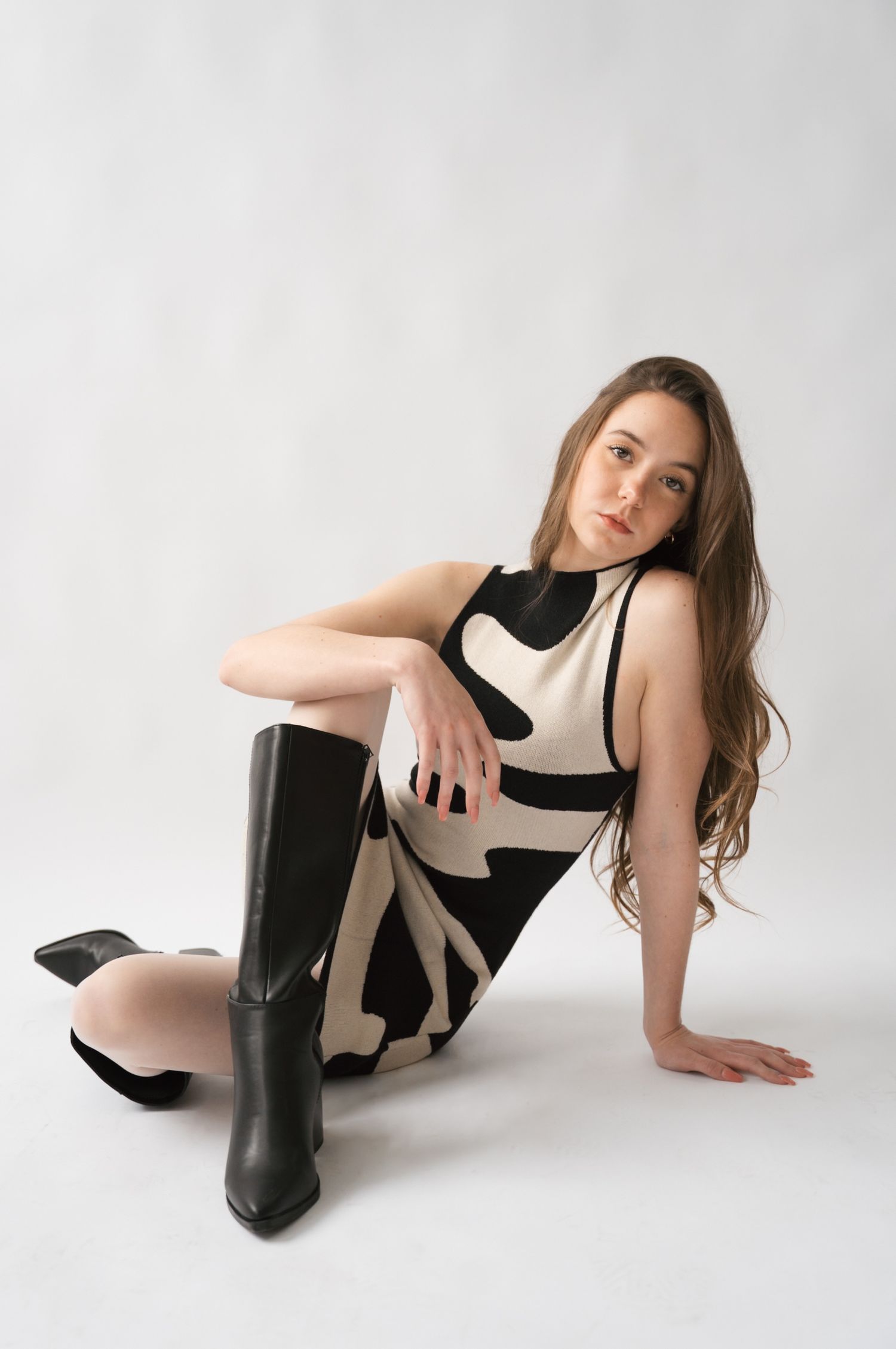 Fashion model poses in black and beige outfit with boots against white background in artistic photo series.