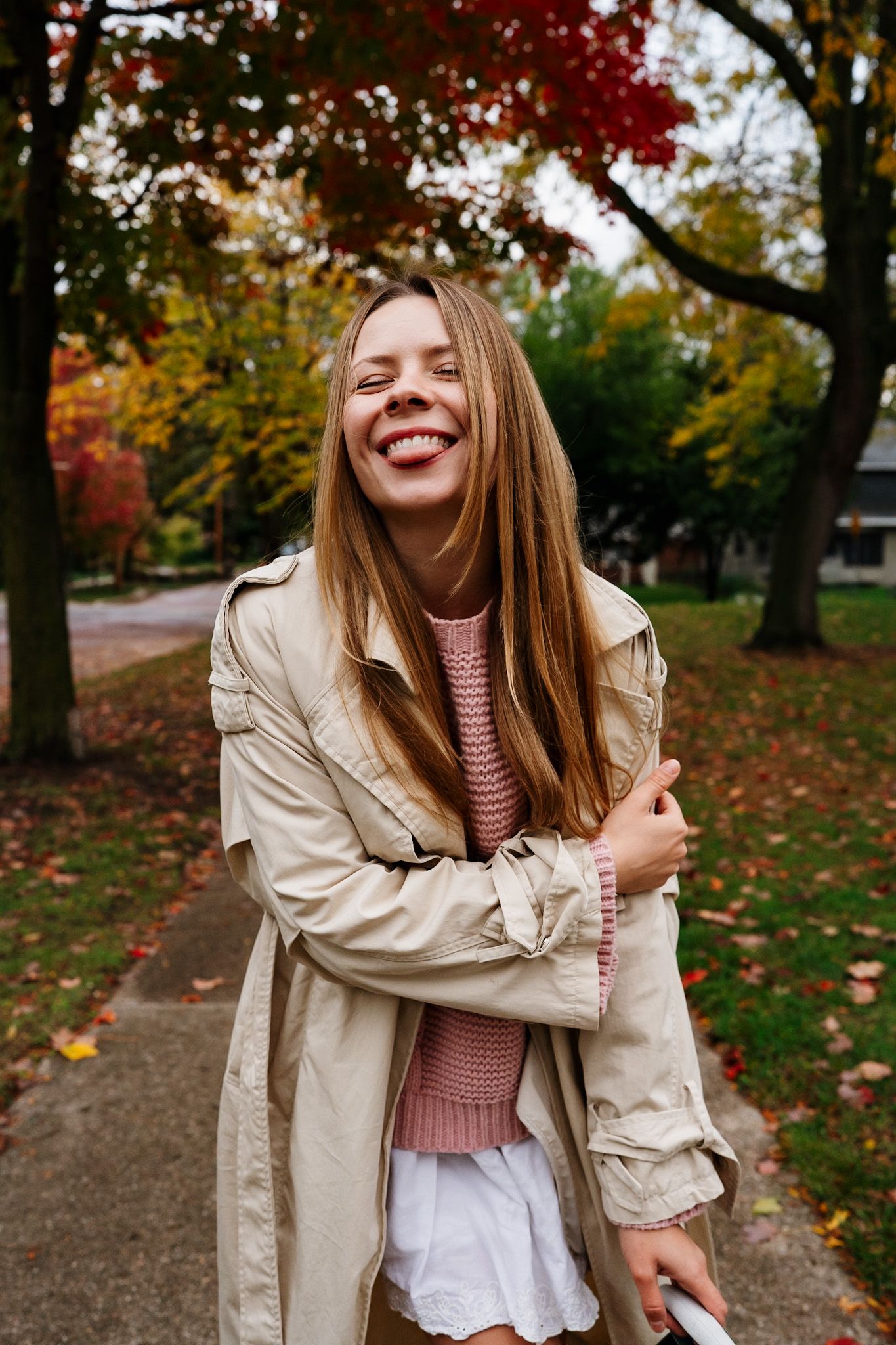 A joyful autumn fashion photoshoot featuring a beige trench coat and pink sweater against colorful fall foliage.