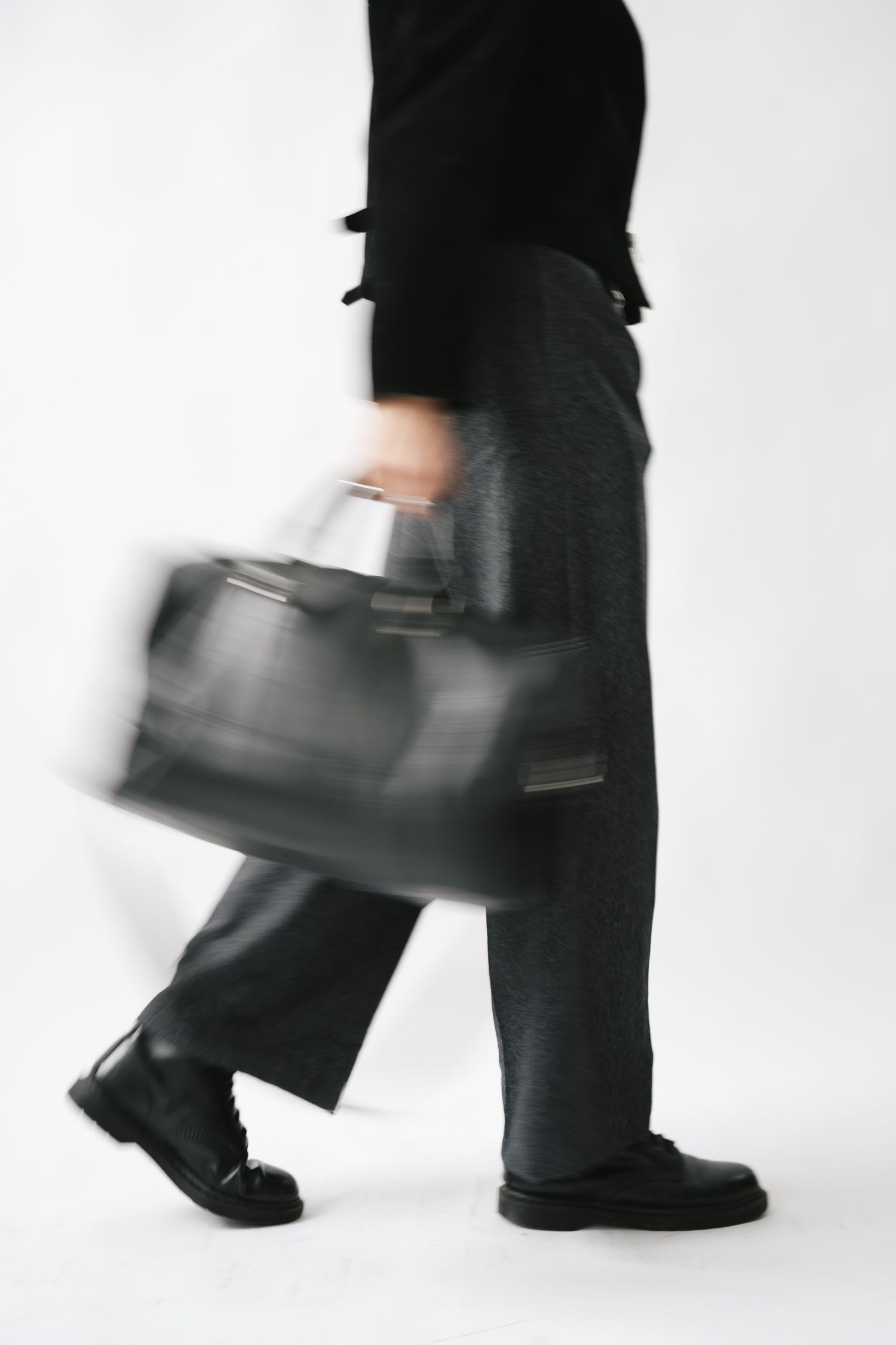 Motion blur photograph of person walking with black leather bag against white background.
