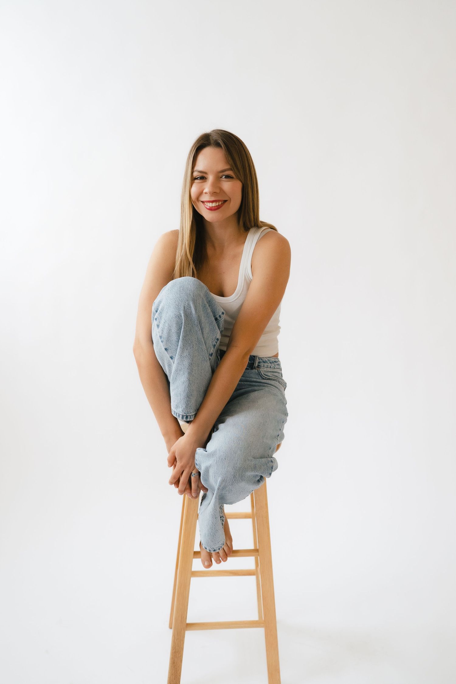 Casual portrait of someone in light denim jeans and white tank top sitting on wooden stool against white background.