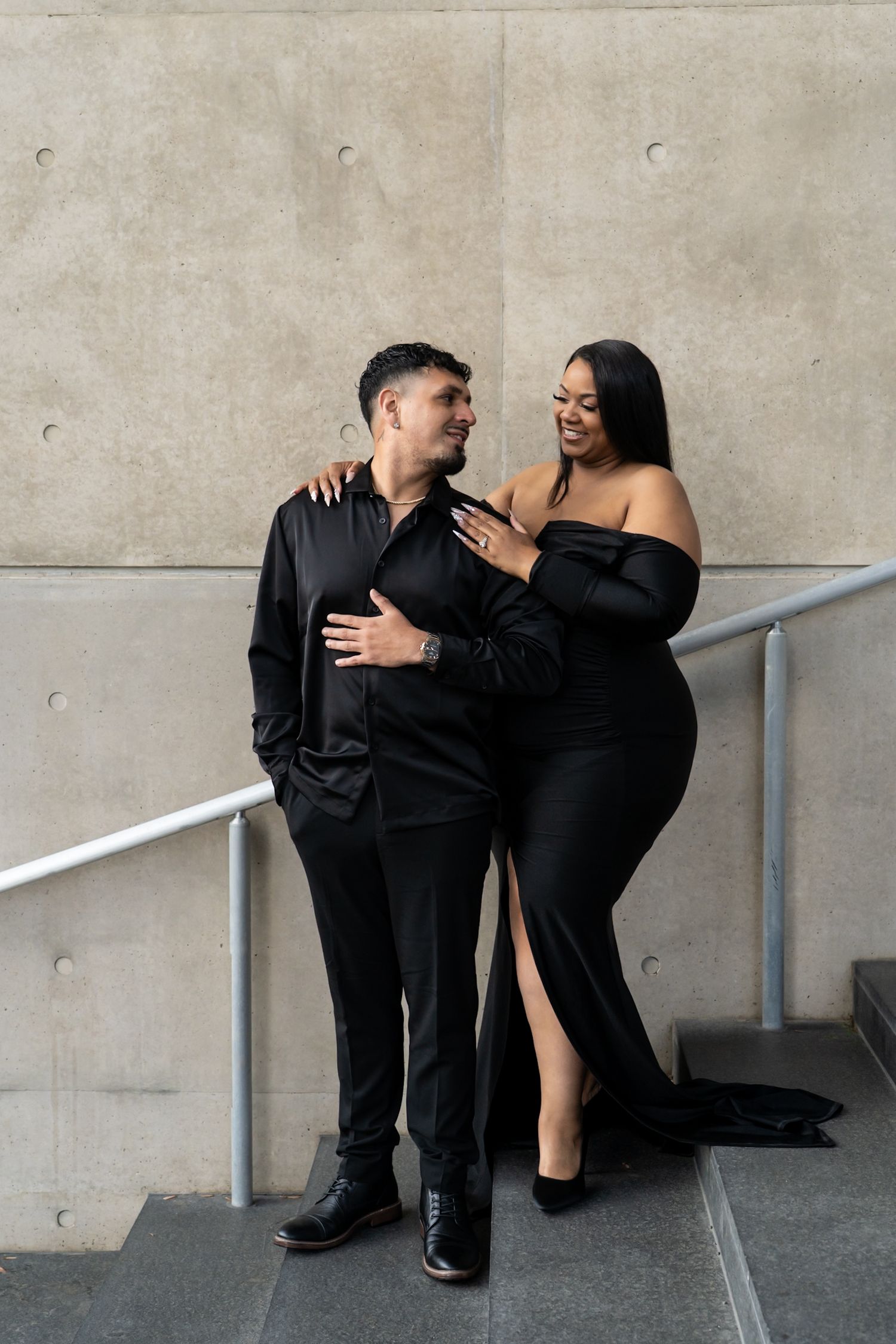 A couple in black formal wear poses playfully against a concrete wall with handrails.