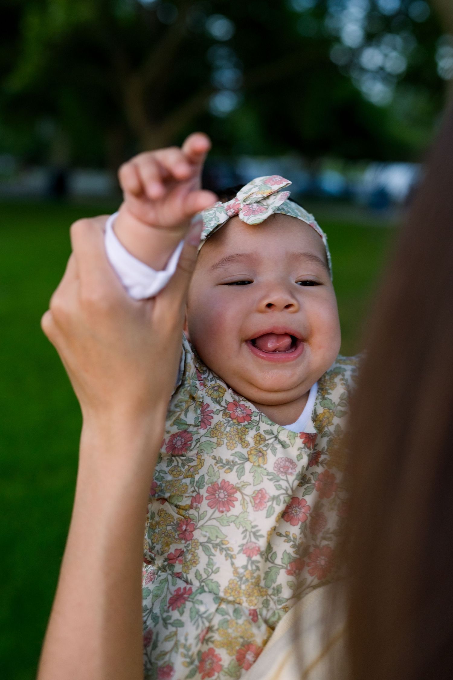 Baby in floral outfit and headband laughs joyfully while being held up outdoors.