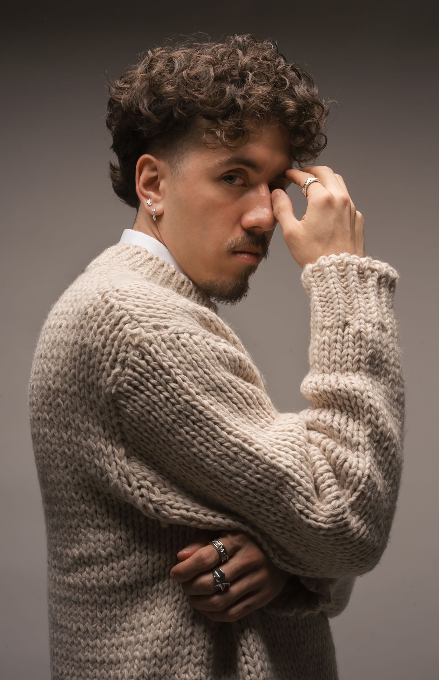 Person in beige knit sweater poses emotively against dark studio background.