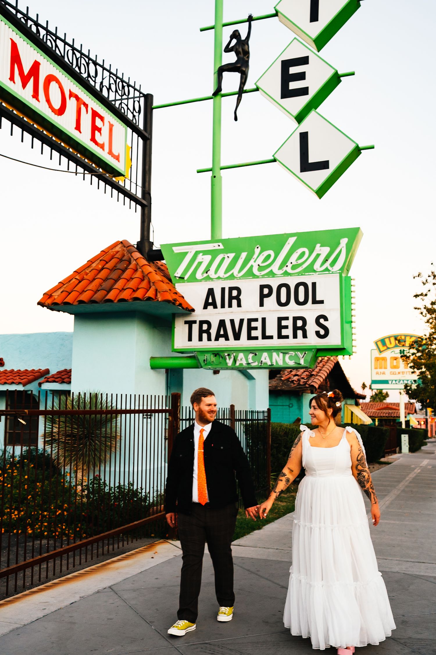 A couple poses outside a retro motel with vintage neon signage for Travelers Air Pool.