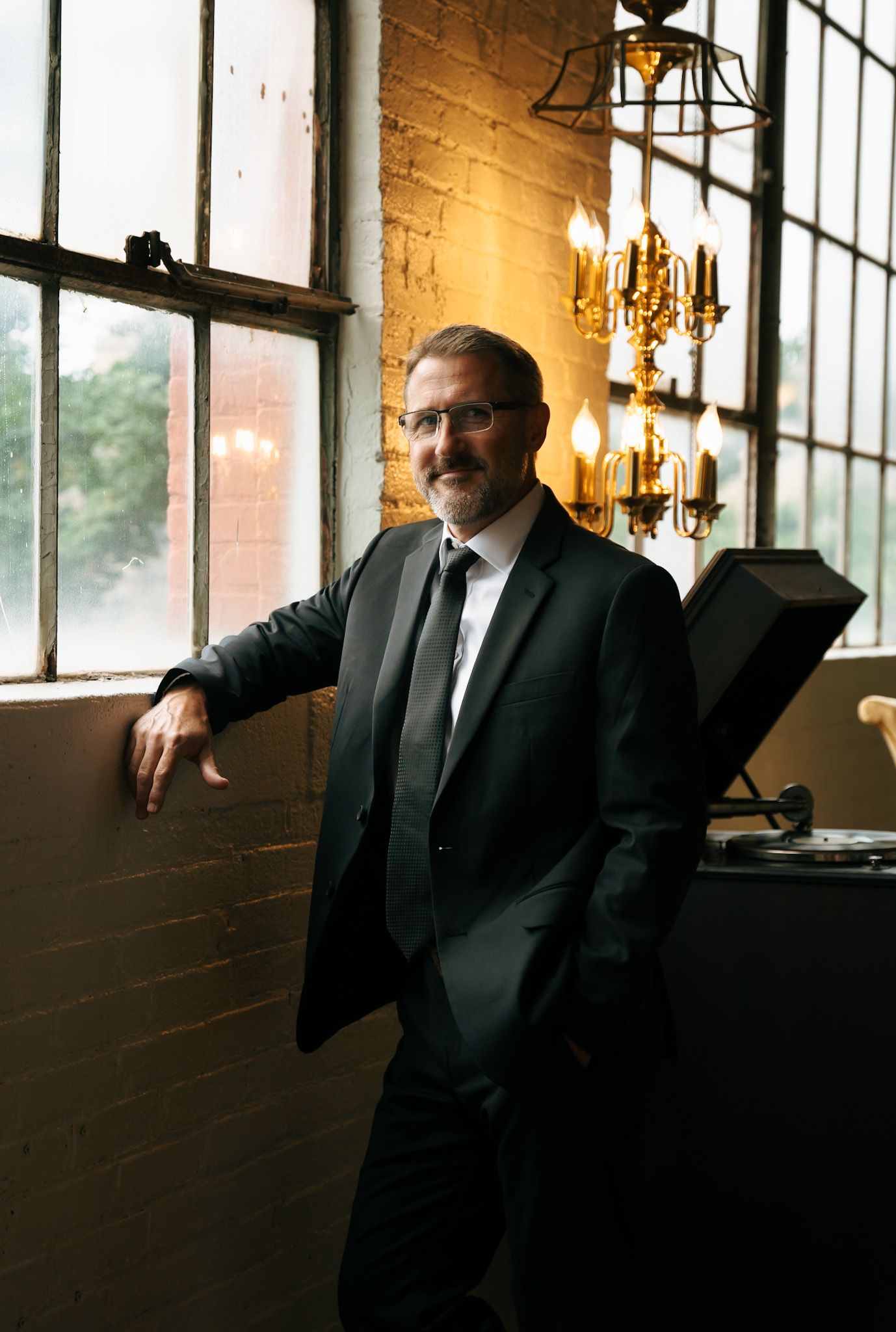 Man in formal attire stands contemplatively by large windows in an elegant indoor setting.