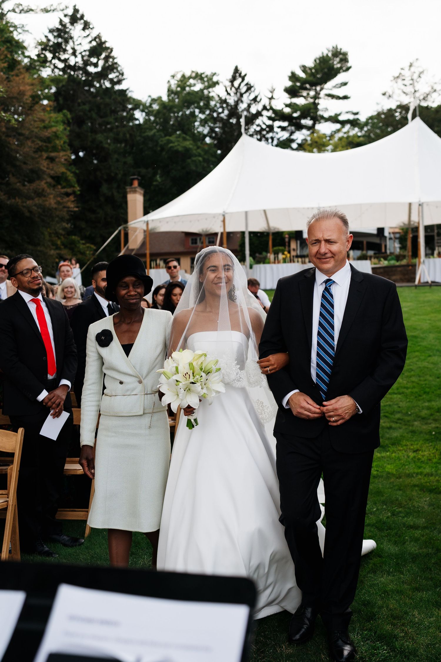 Wedding processional on lawn with white tent and wooden chairs in background.