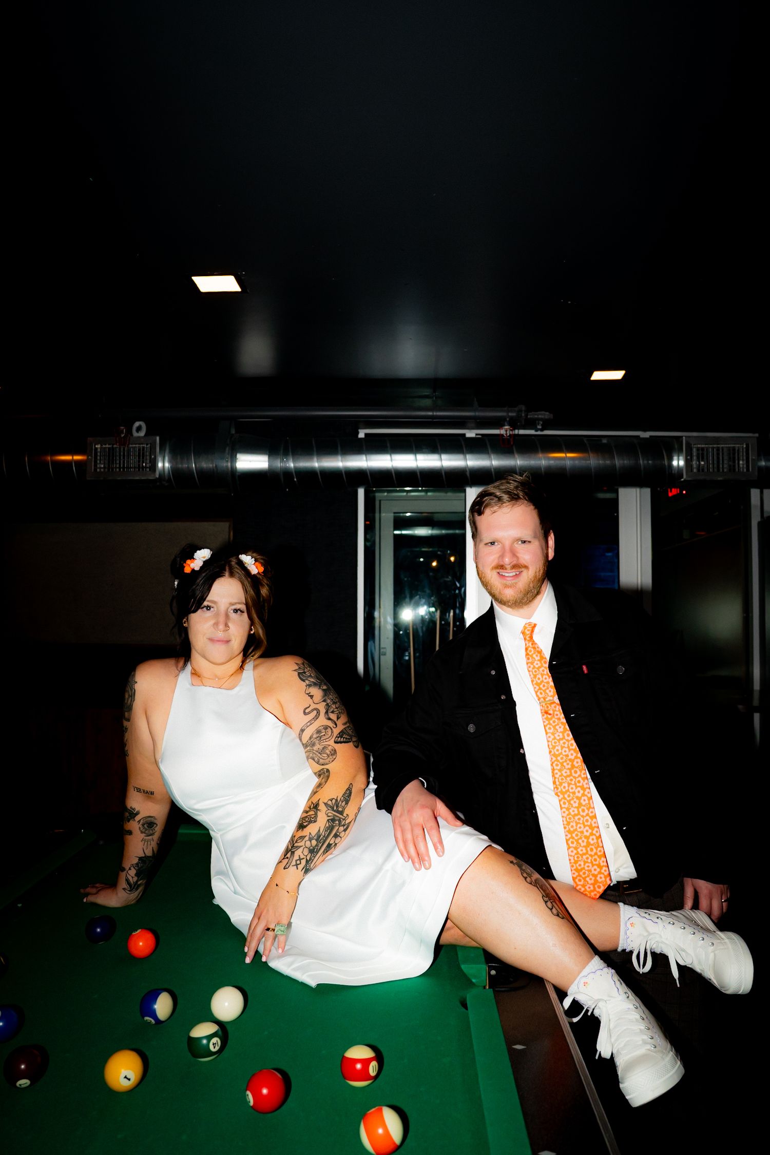 A playful moment on a pool table with a couple in wedding attire, showing the bride sitting on the table's edge.