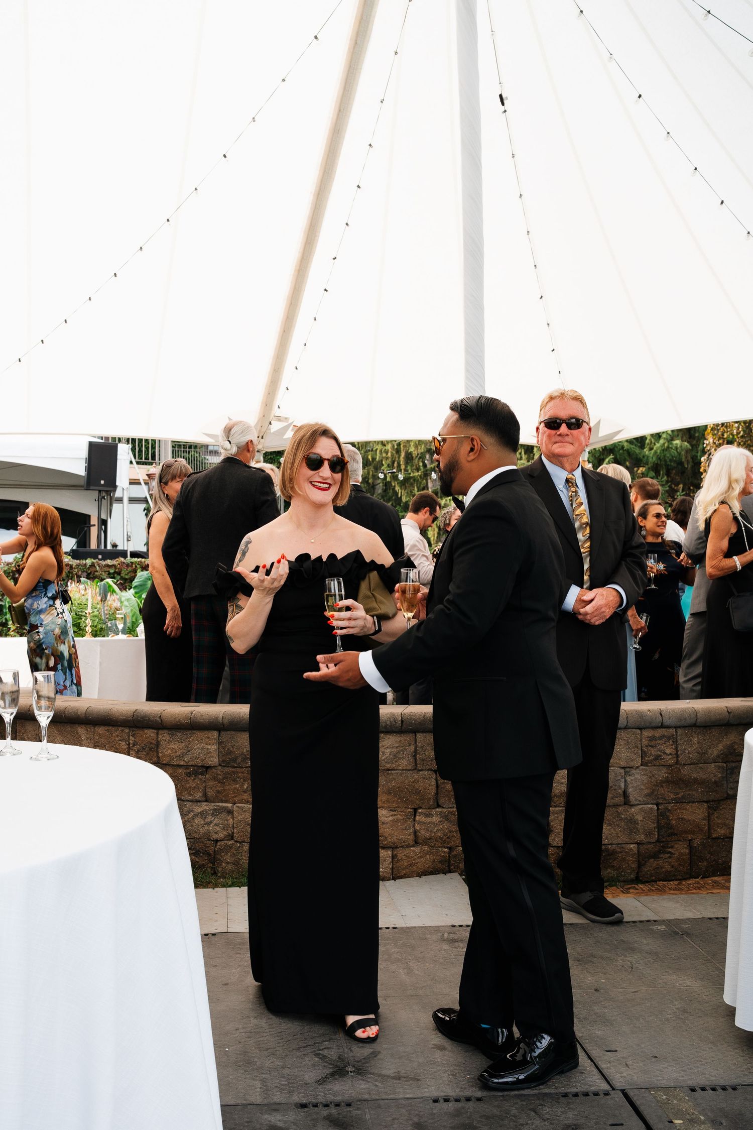 Wedding reception under a white tent with guests mingling near stone wall and cocktail tables.