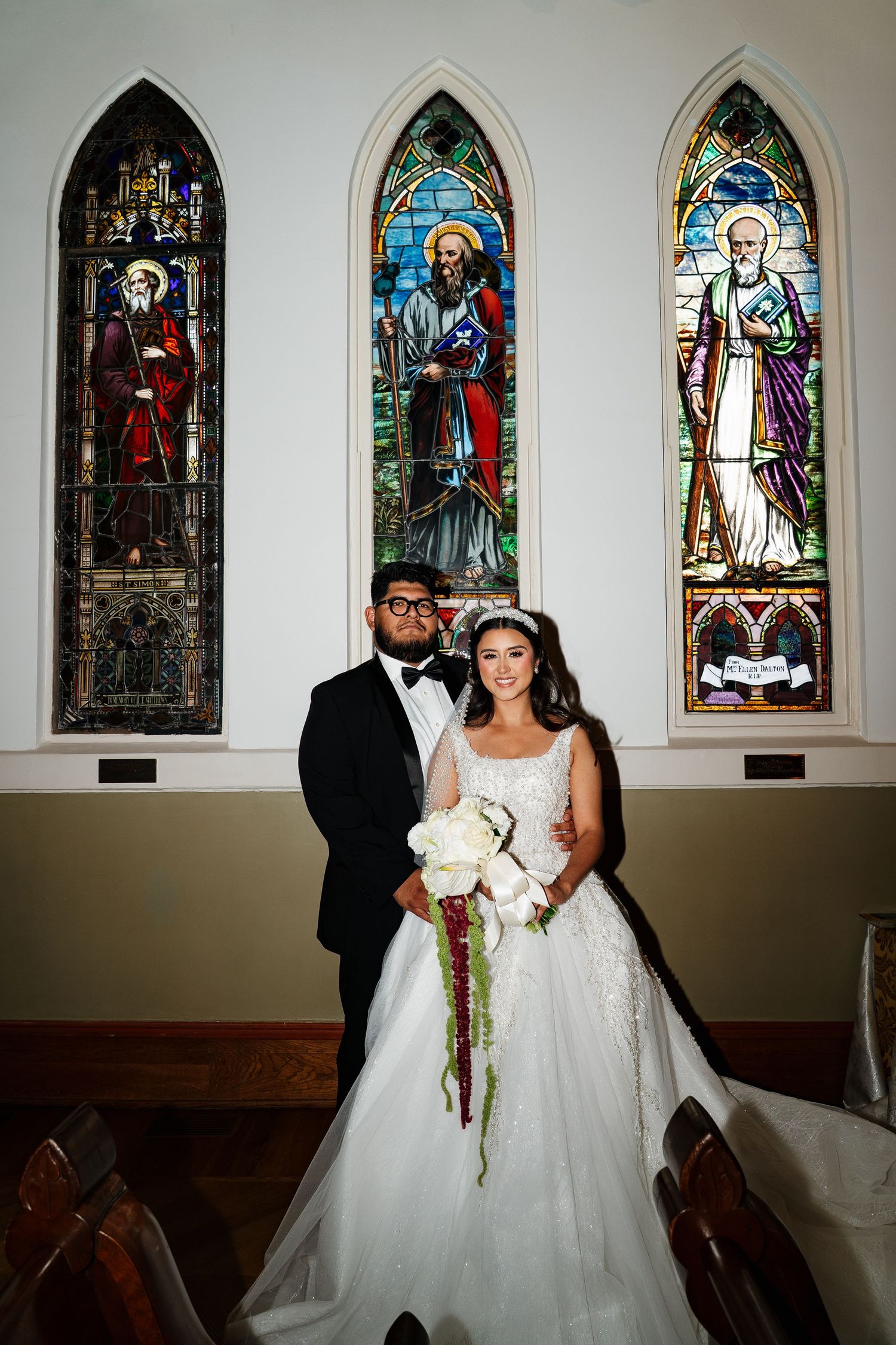 A couple in wedding attire standing in front of stained glass church windows.
