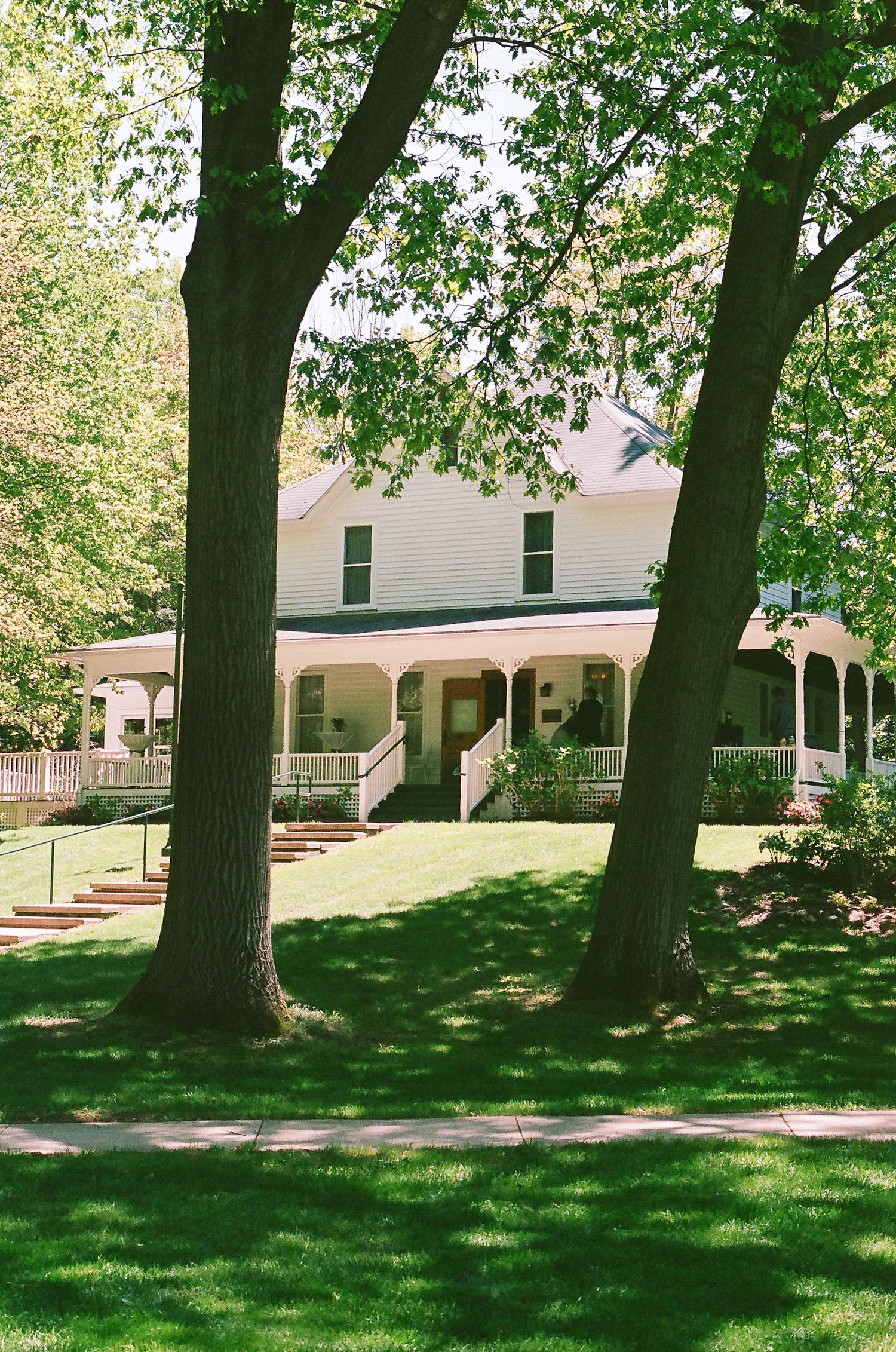 White farmhouse with wraparound porch nestled among mature trees on green lawn.