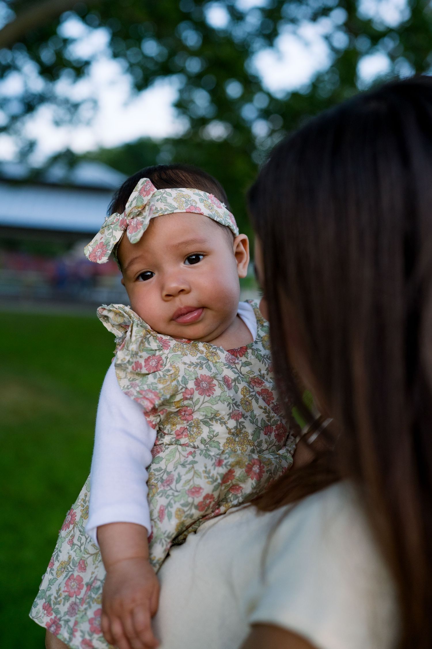 Baby wearing floral dress and matching headband looks over caregiver's shoulder in outdoor setting.