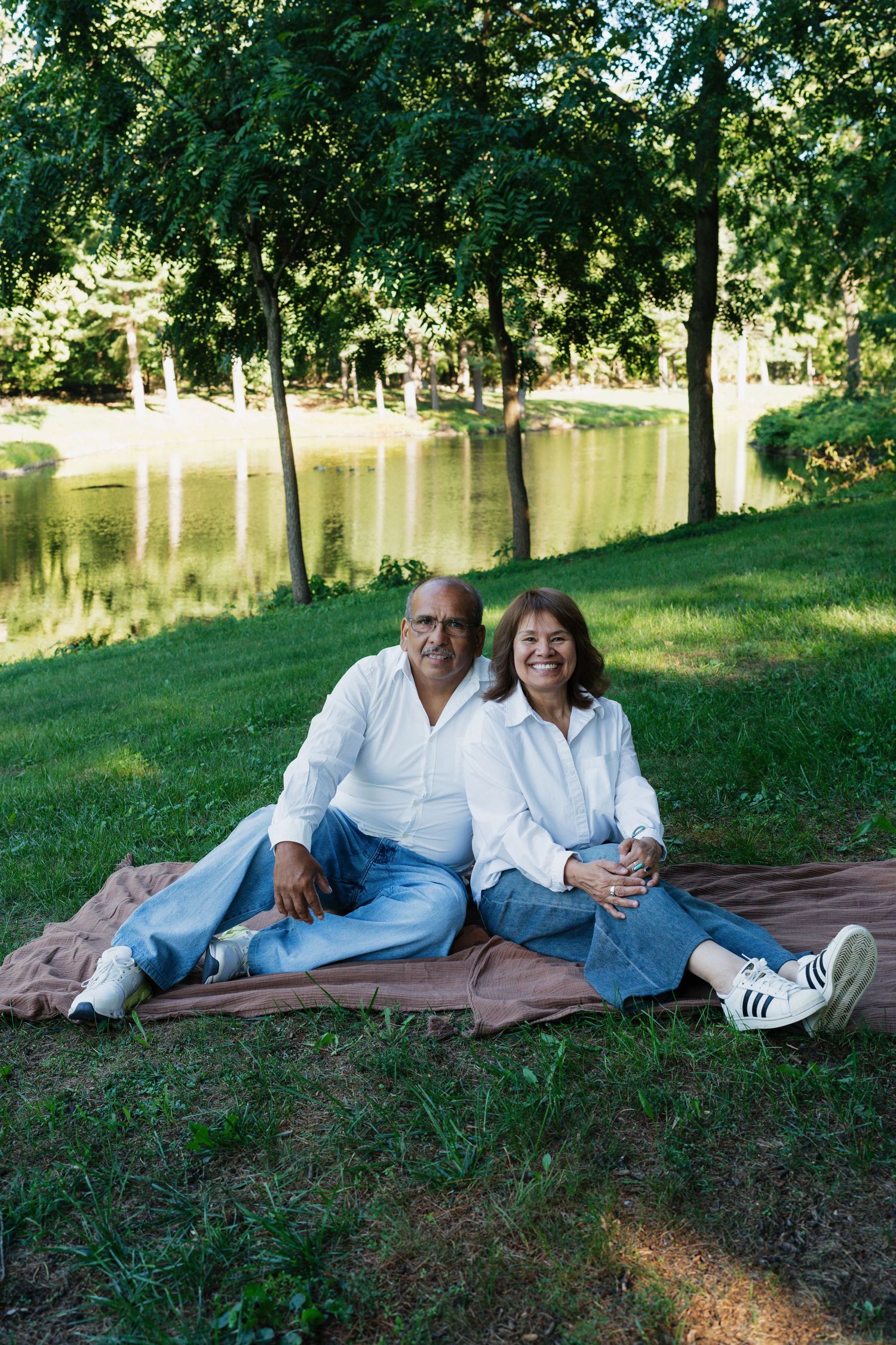 Couple relaxing on blanket by lake under green trees on sunny afternoon.