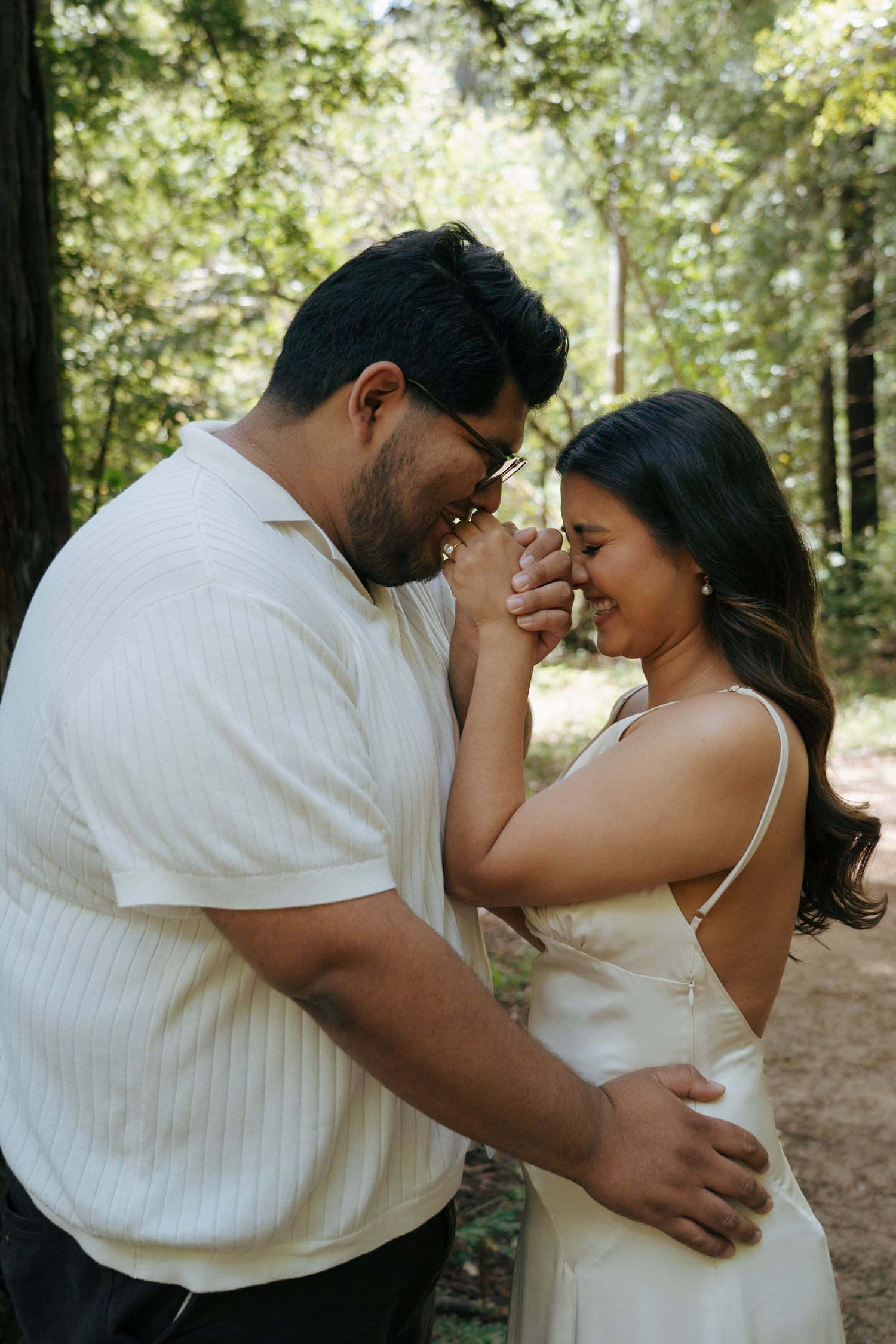A couple shares an intimate forehead touch moment in a sunlit forest setting.