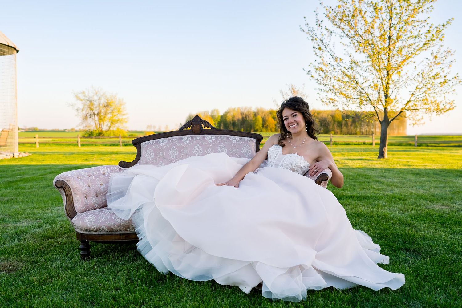 A flowing white wedding dress spreads elegantly across a vintage sofa in an outdoor setting.