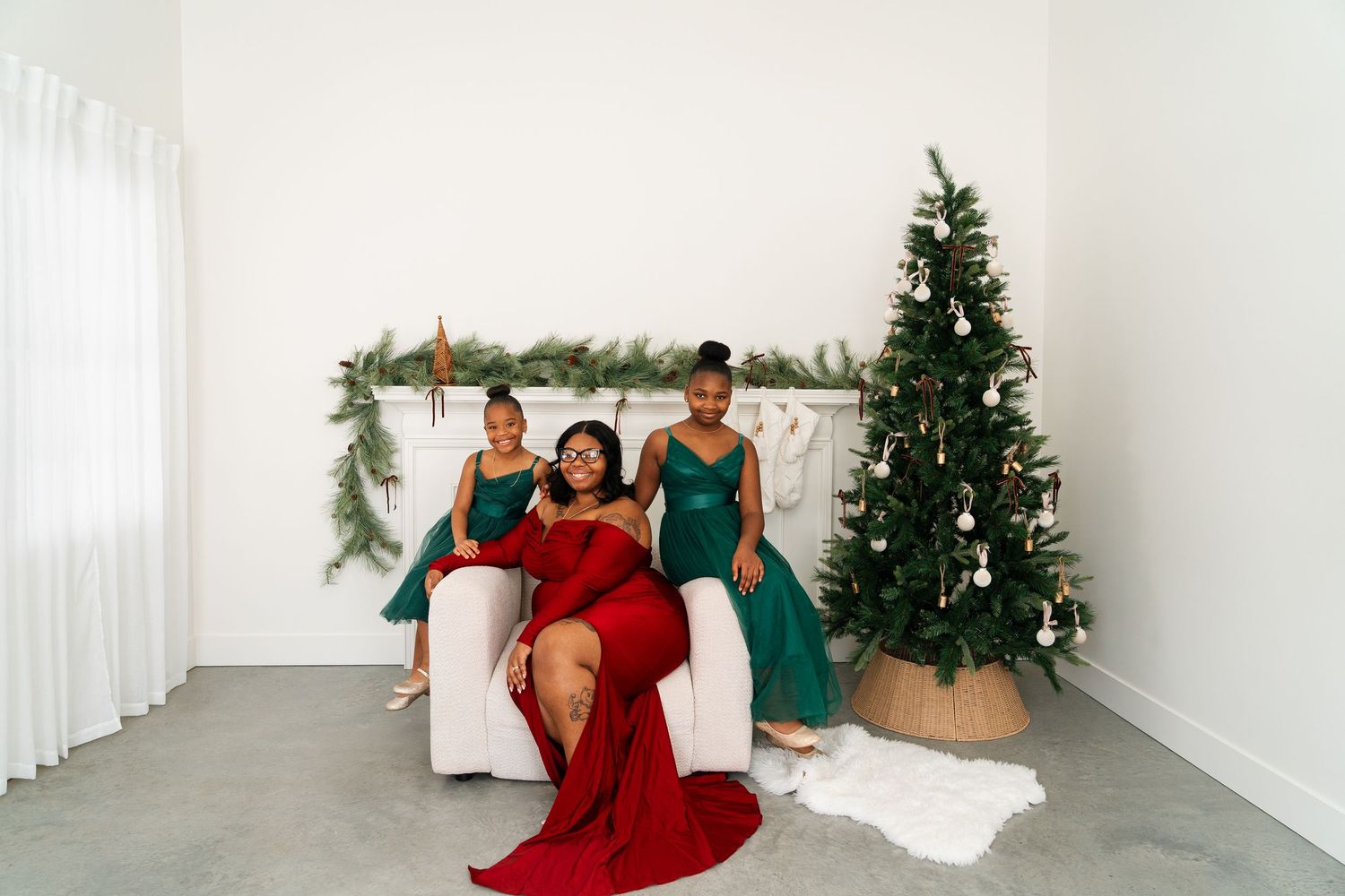 Three women in elegant dresses pose together in a festive holiday setting with a Christmas tree and garland.