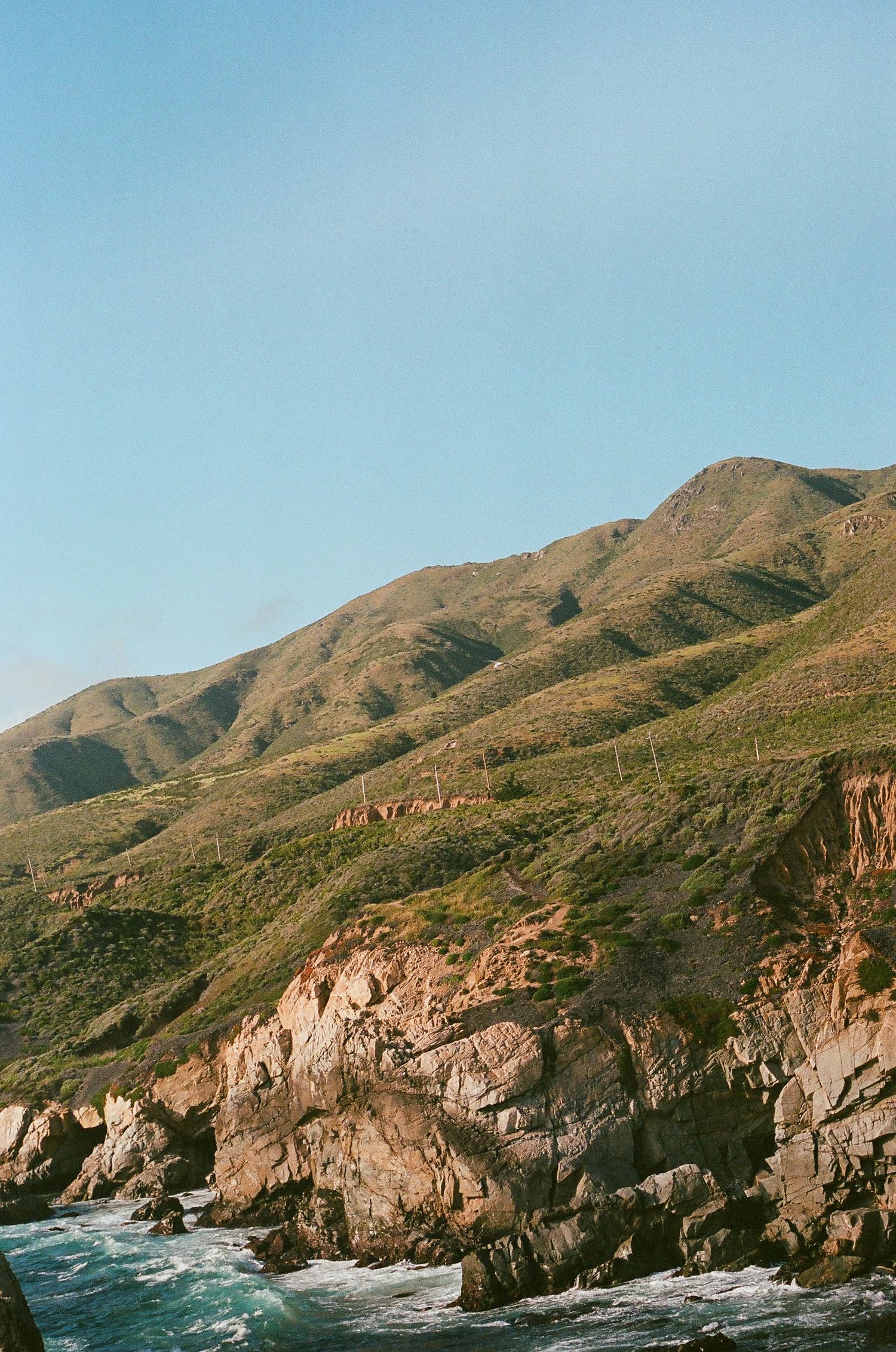 Rocky coastal cliffs and mountains meet crashing waves along a rugged shoreline under clear blue skies.