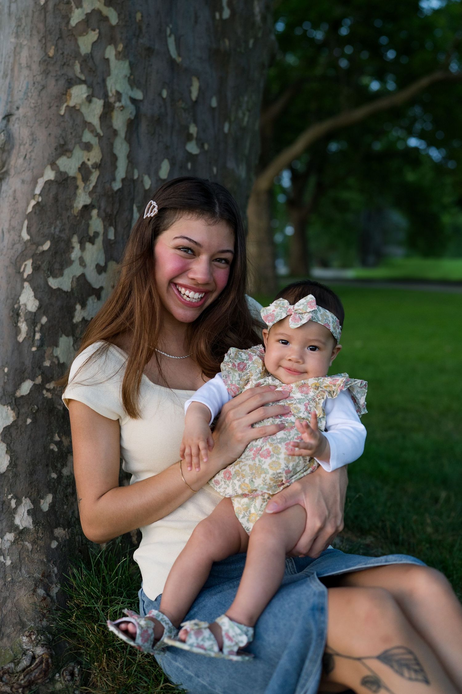 Parent and baby in floral dress share joyful moments under shady tree.
