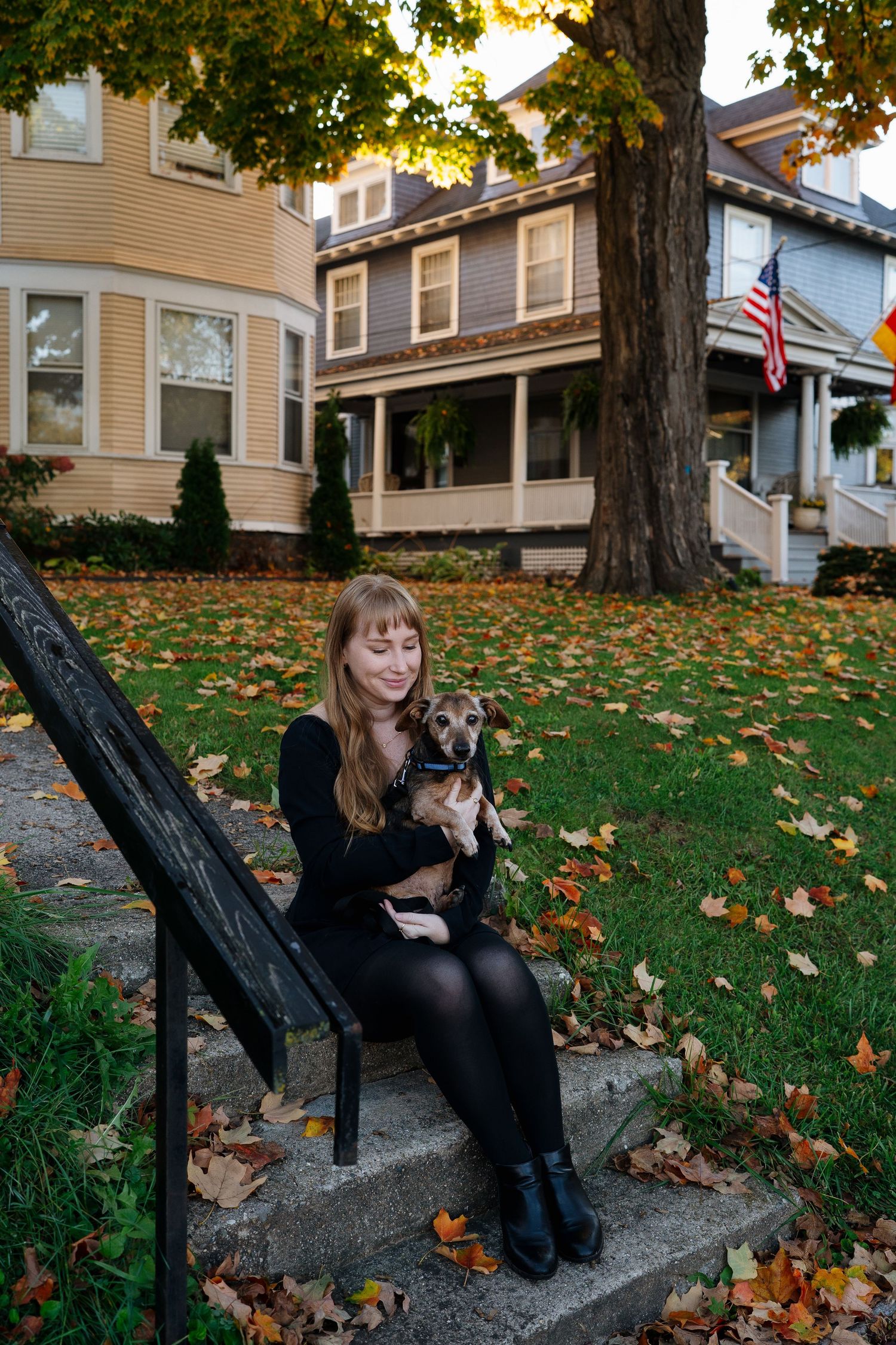 Person sits on steps with small dog as fallen leaves scatter around historic homes.