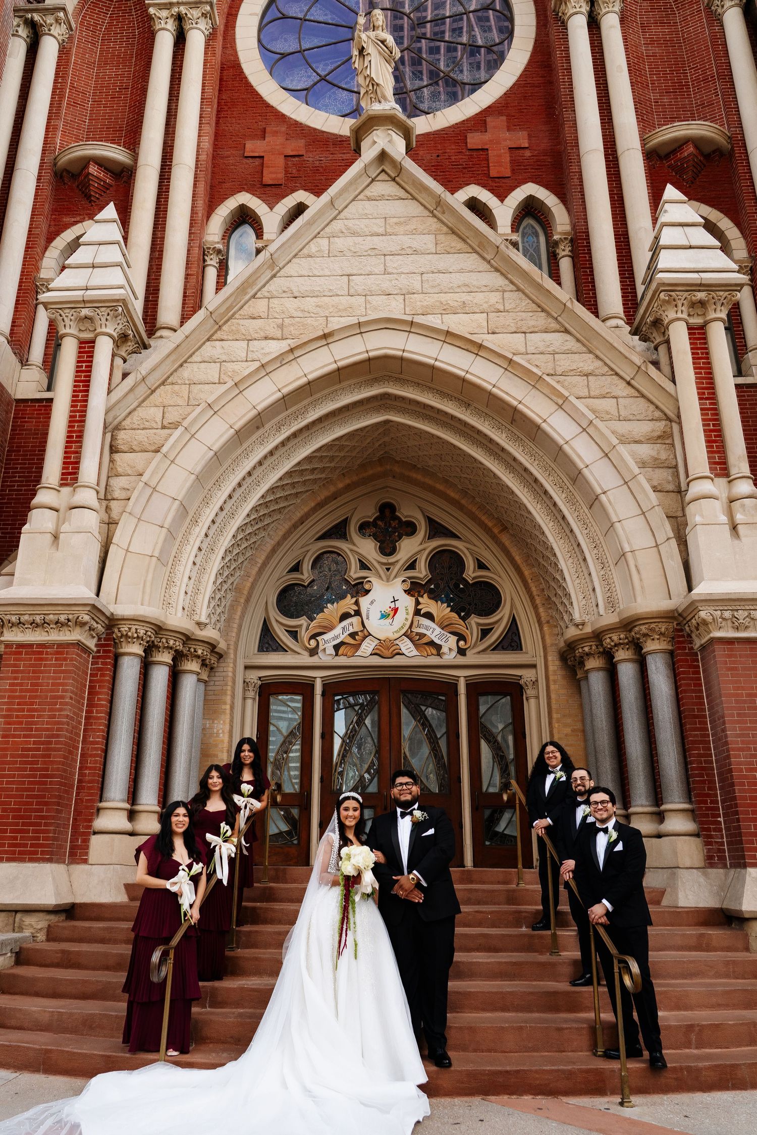 Wedding ceremony group photo outside ornate cathedral entrance with burgundy and black attire.