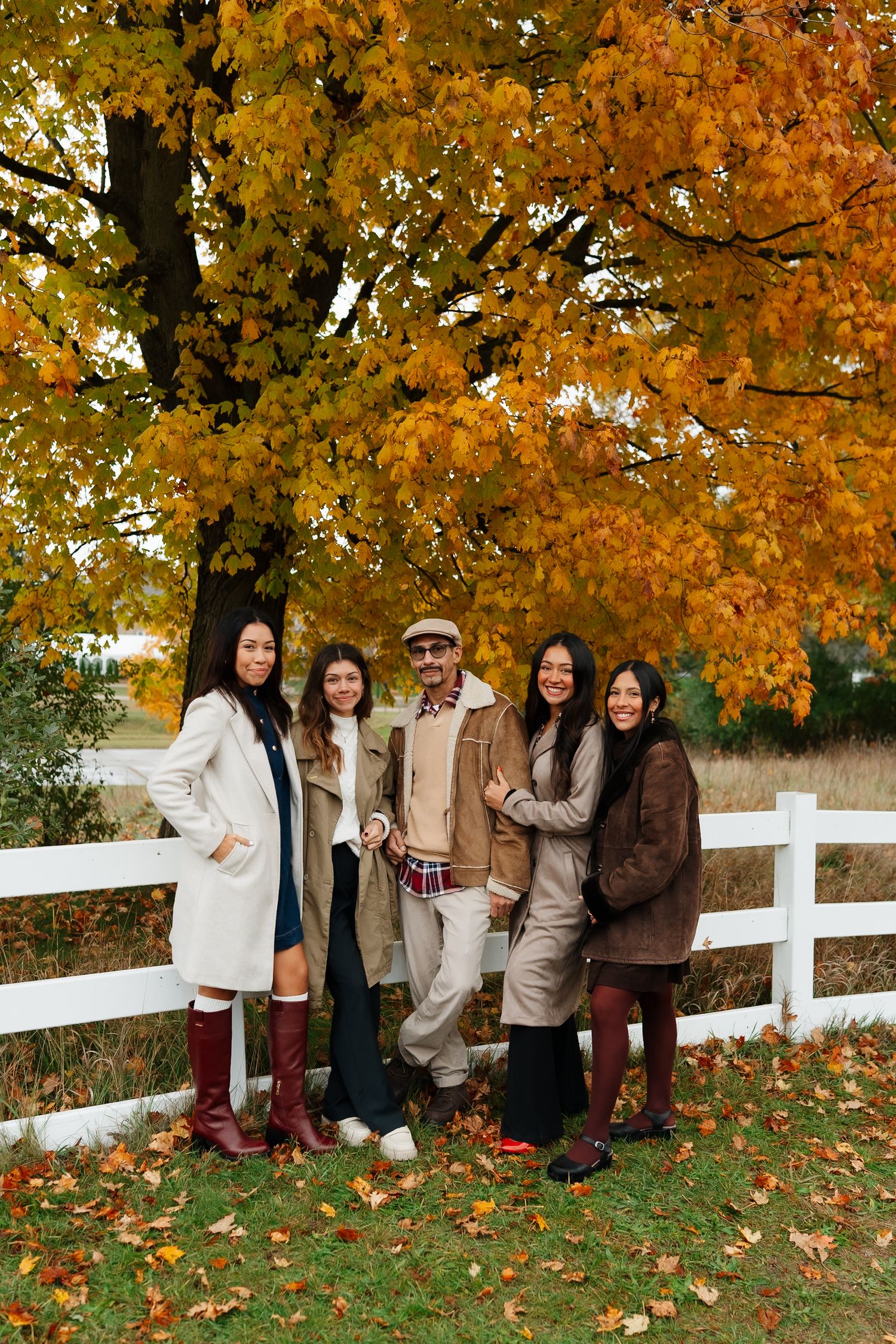 Family portrait beneath golden autumn tree with white fence in foreground.