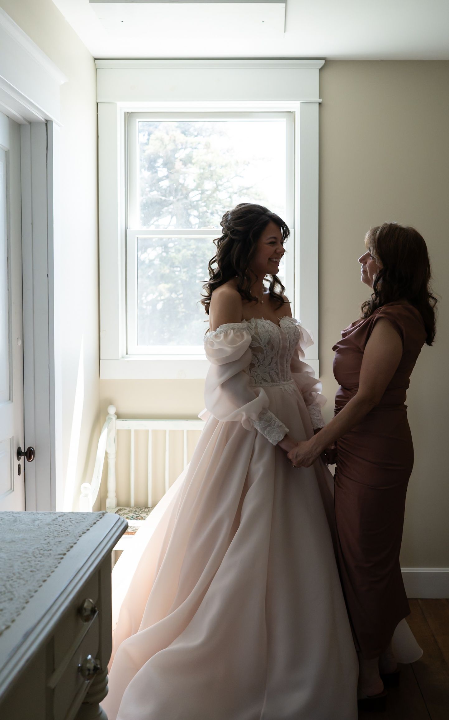 Intimate getting ready moment as helpers assist with a wedding dress in natural window light.