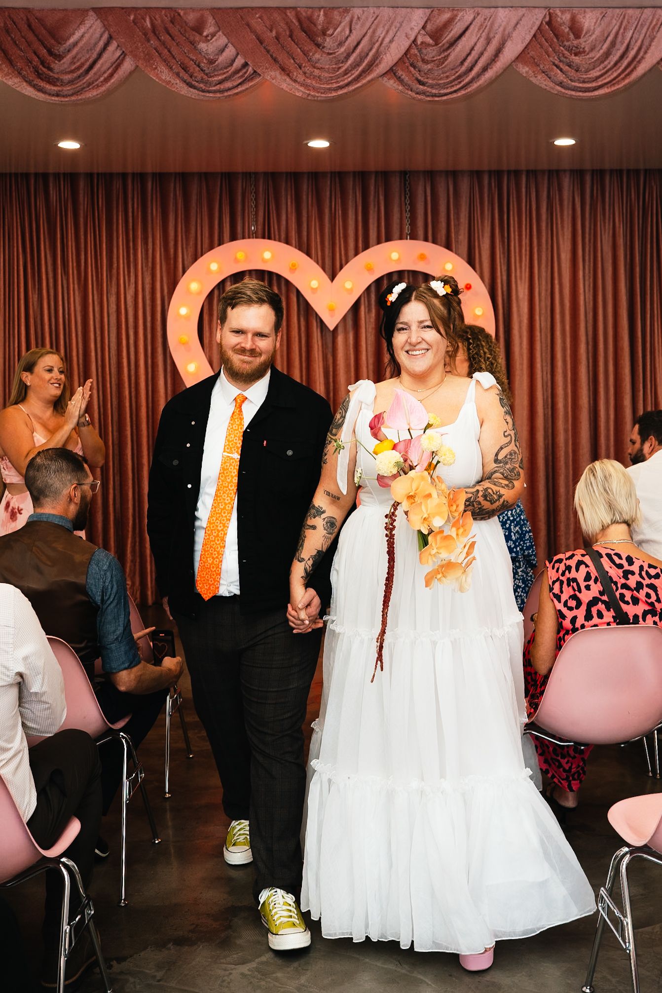 A wedding couple walking under a heart arch with the bride in a white gown and groom in black suit.