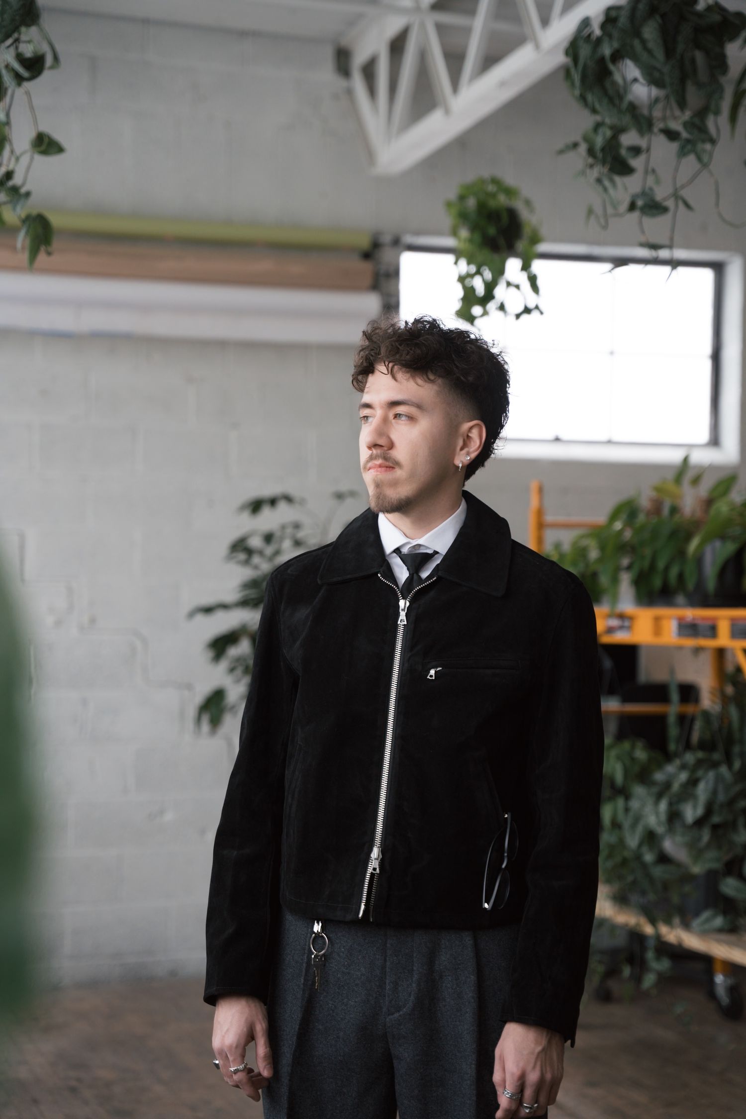 Model in black jacket poses in industrial space with plants and white walls.