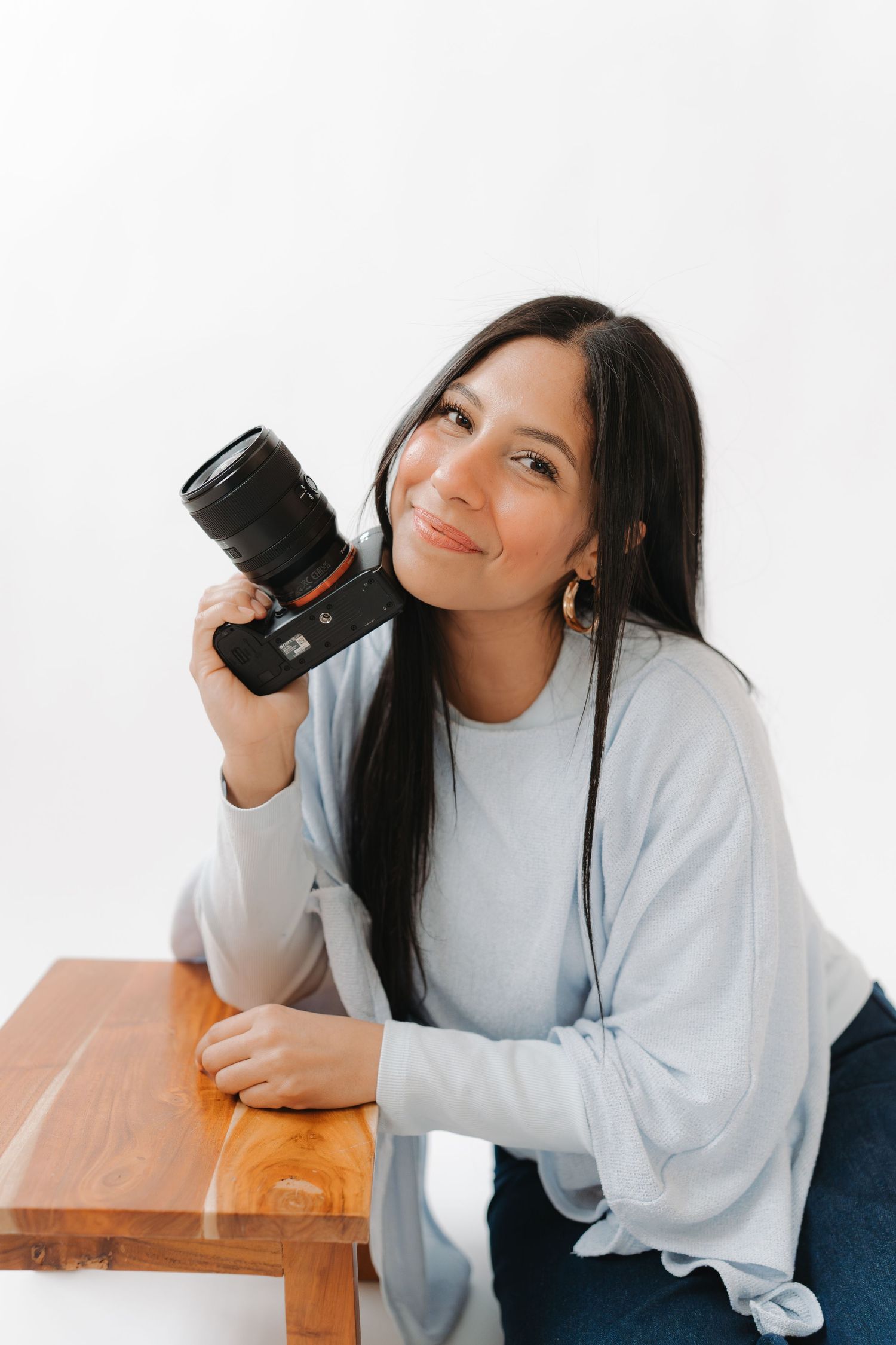 A person in a white sweatshirt poses playfully with long dark hair while sitting at a wooden table.
