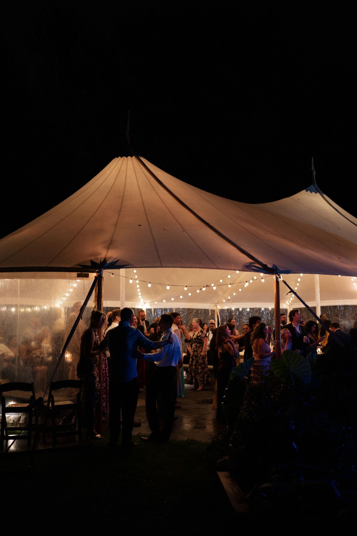 Wedding reception tent illuminated with string lights as guests dance and celebrate inside at night.