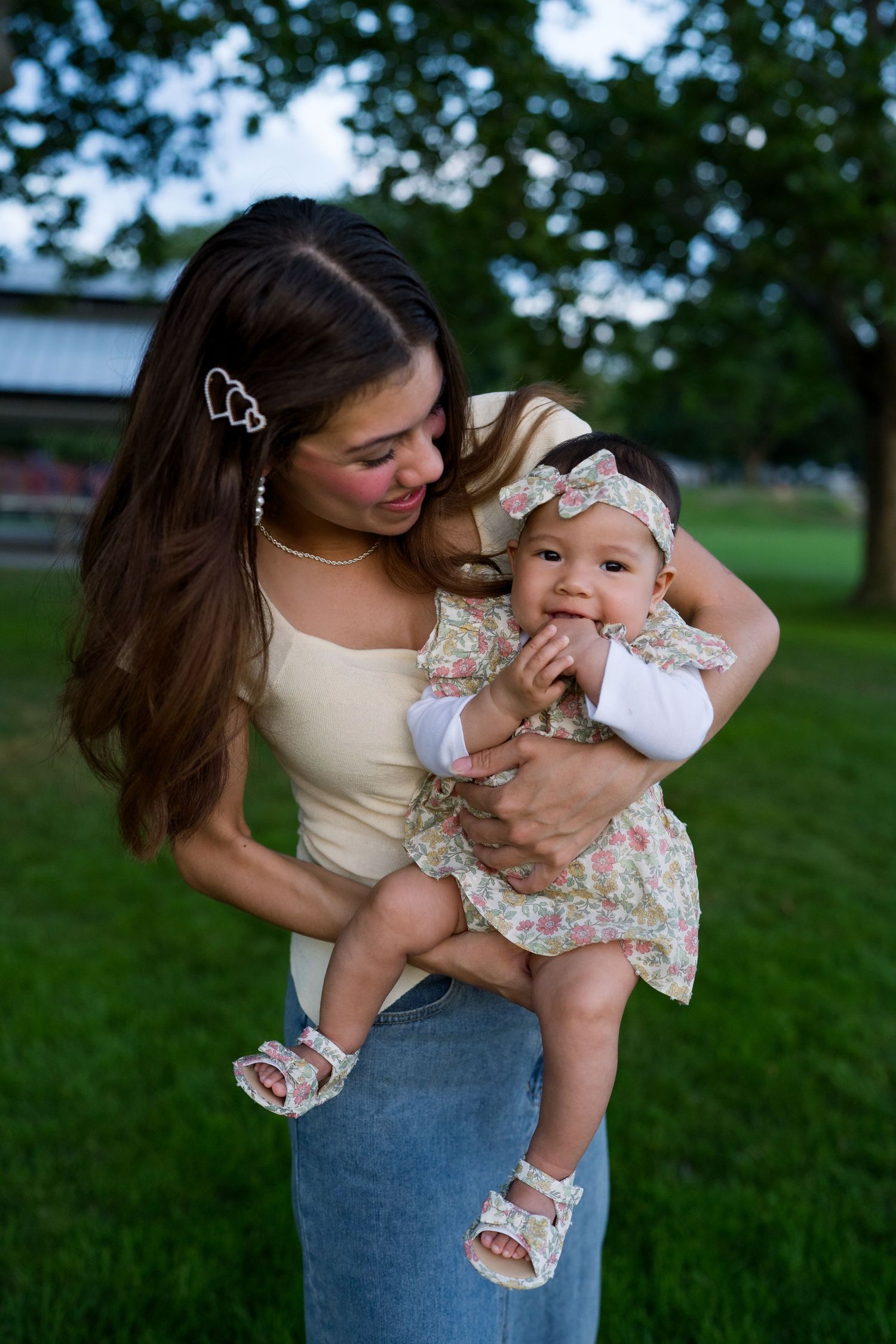 Parent holding baby in floral outfit during outdoor photoshoot in park setting.