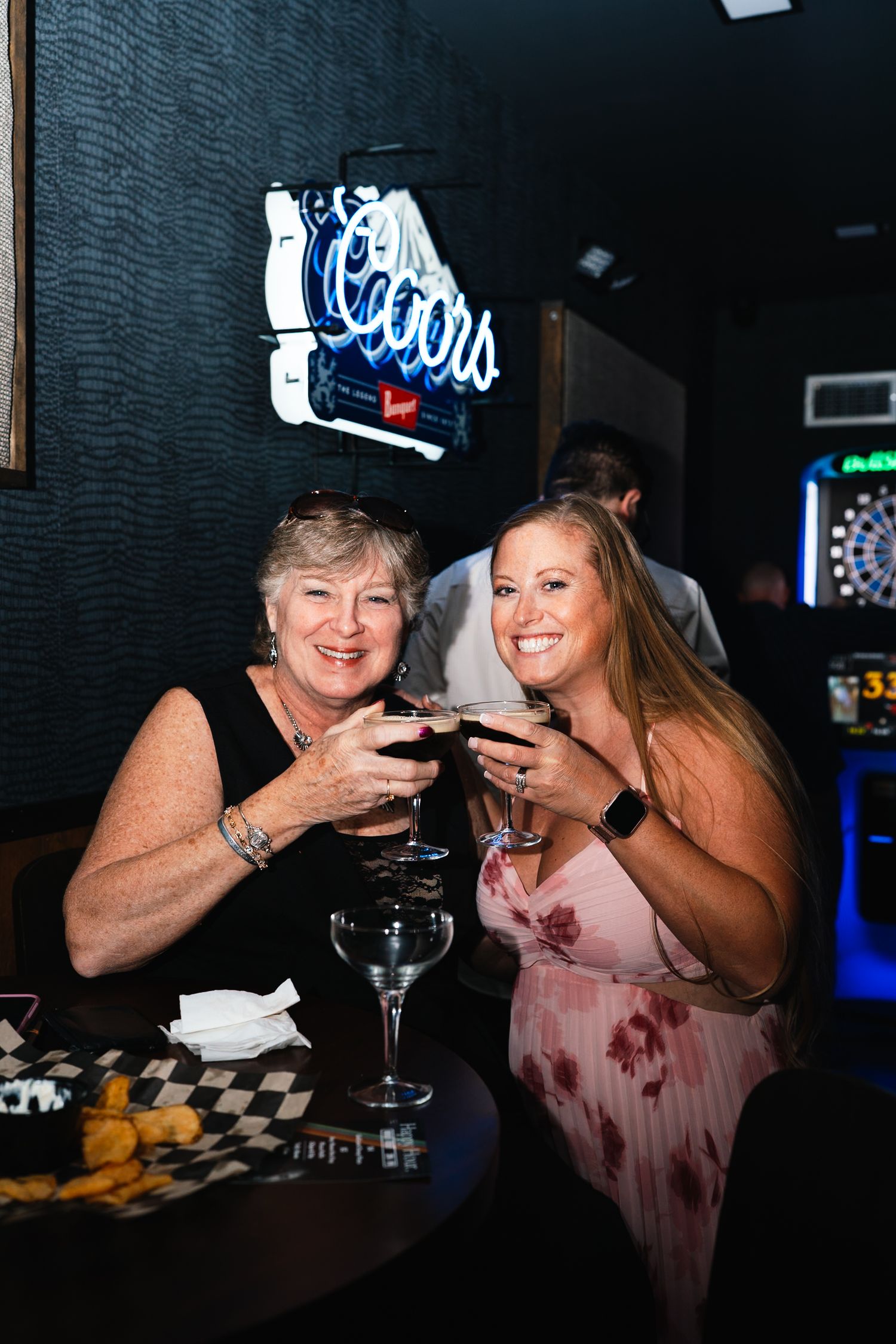 Two people enjoying drinks together under a neon Coors sign in a dimly lit bar.