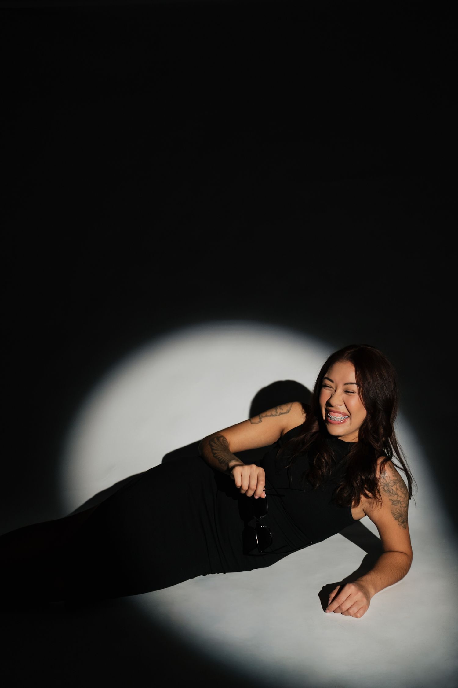Dramatic studio portrait with person in black dress lying in spotlight against dark background.