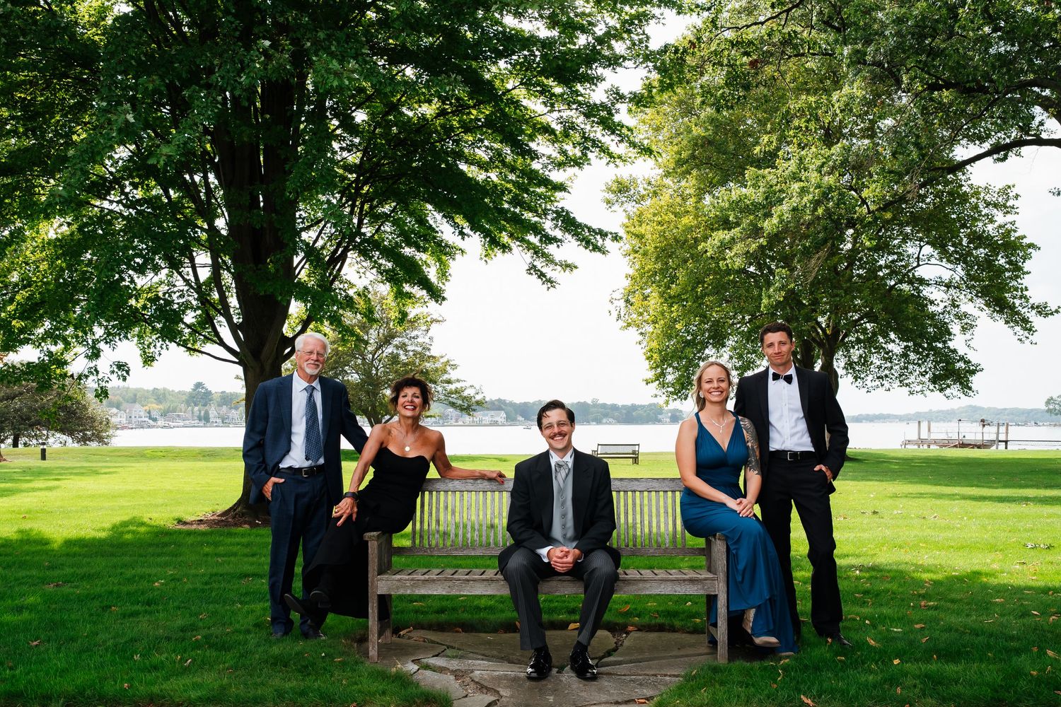 Wedding party relaxes on a grassy lawn with large trees in the background.