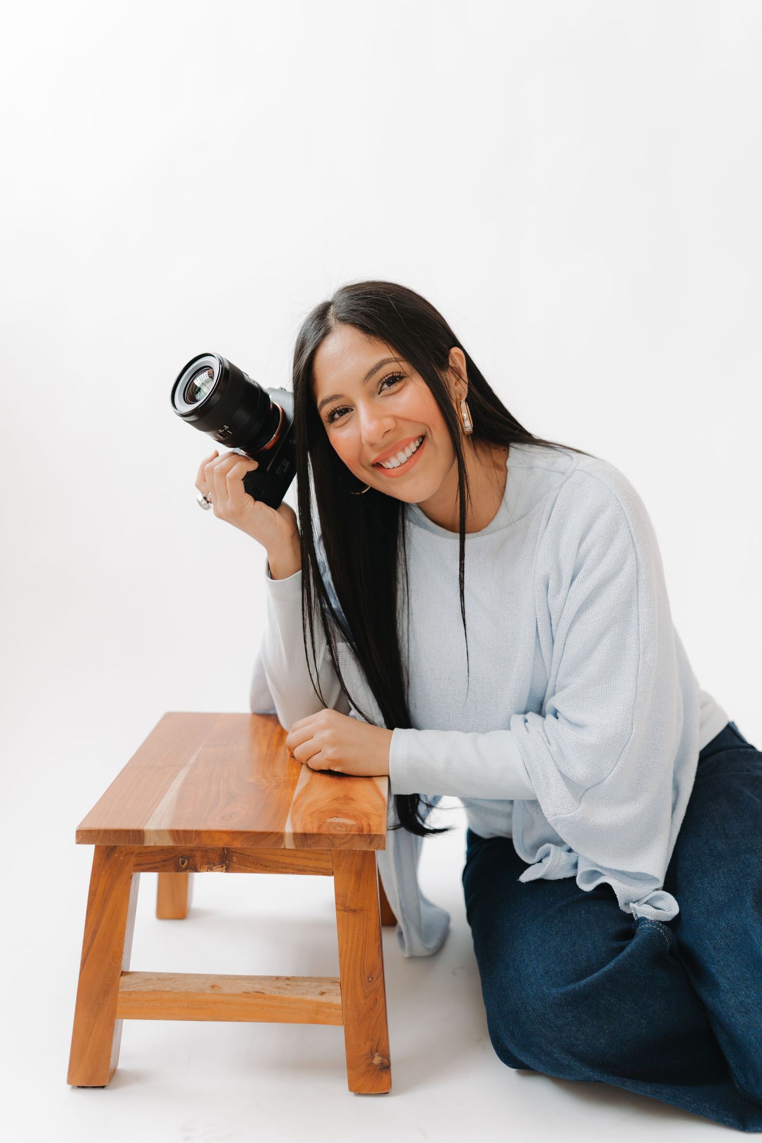 A person in a white sweater and jeans poses playfully with a wooden stool in a photo series against a white backdrop.