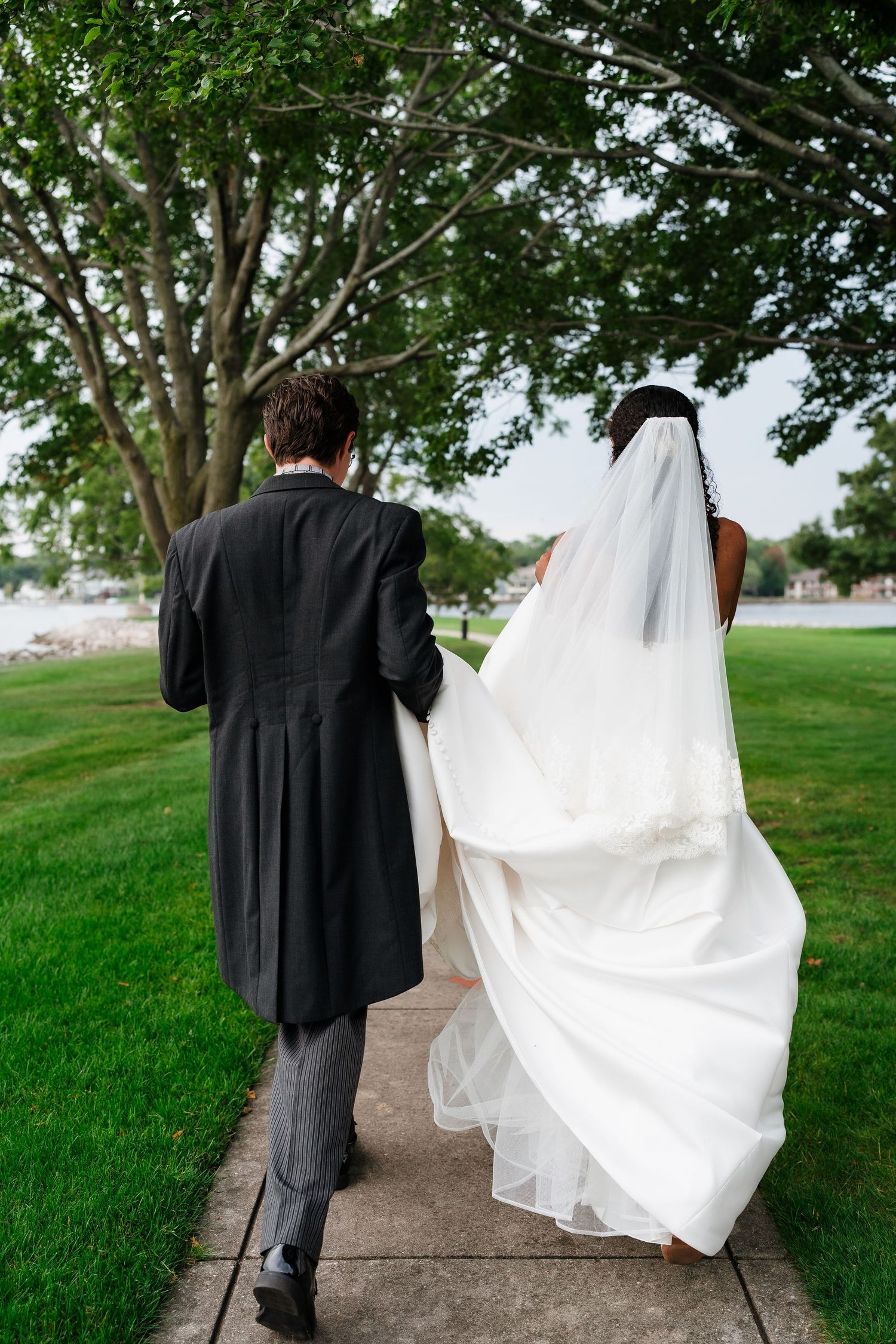 A couple walks along a path outdoors with the bride's white dress flowing in the breeze.