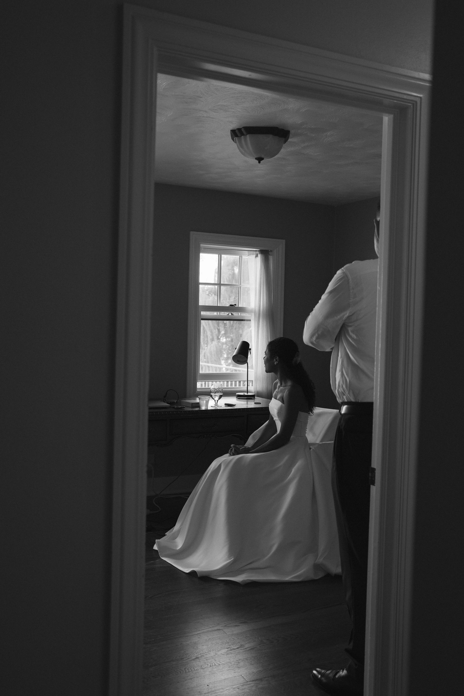 Black and white photo through doorway showing bride sitting by window in elegant dress.
