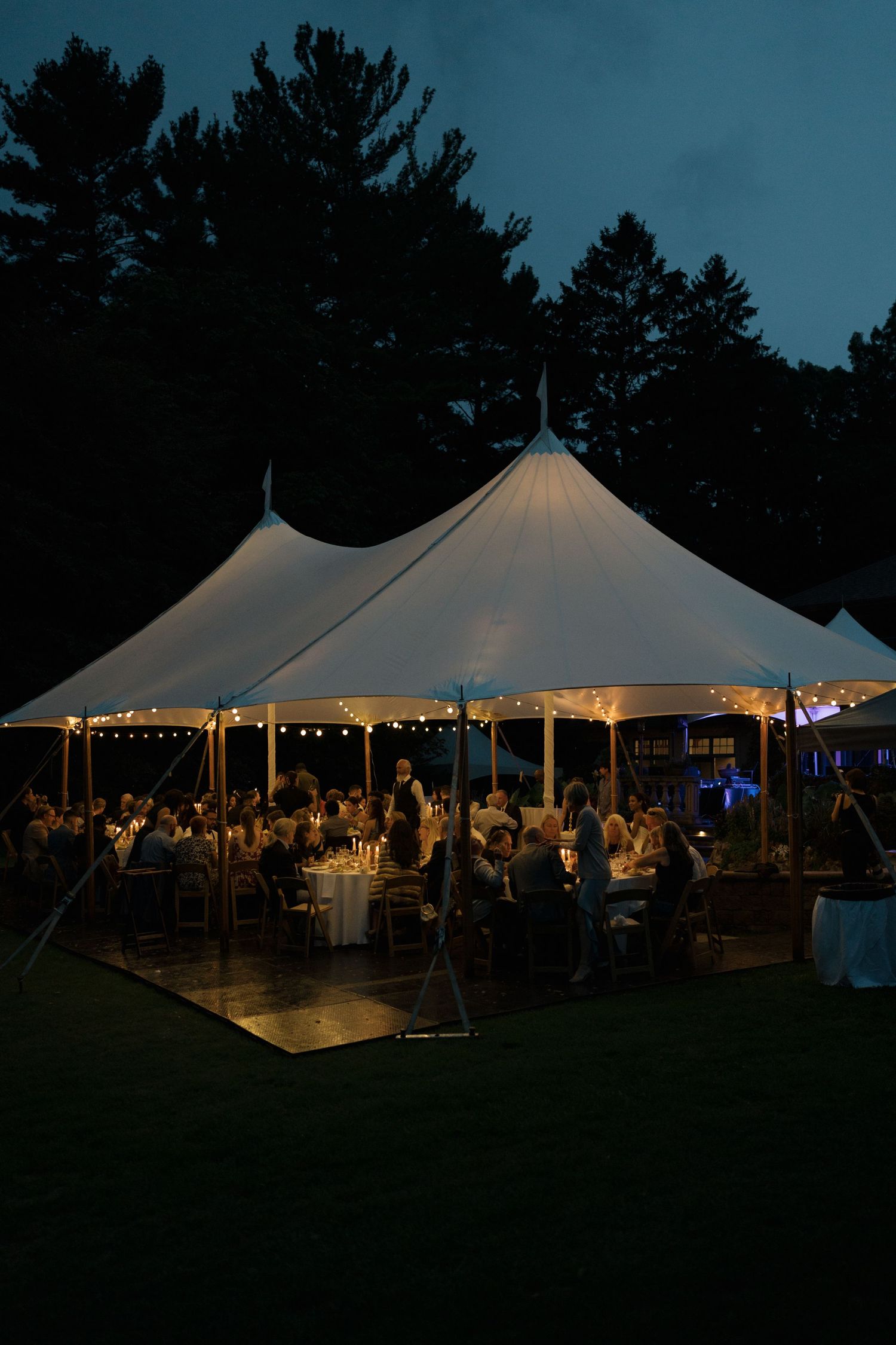 Large white wedding reception tent illuminated at dusk with string lights.