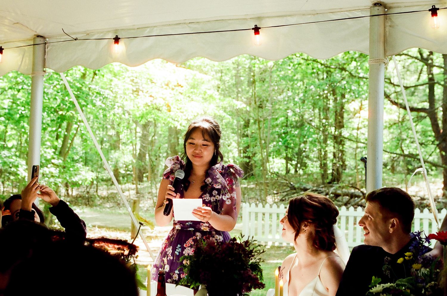 A festive outdoor reception under a white tent with string lights and greenery in the background.