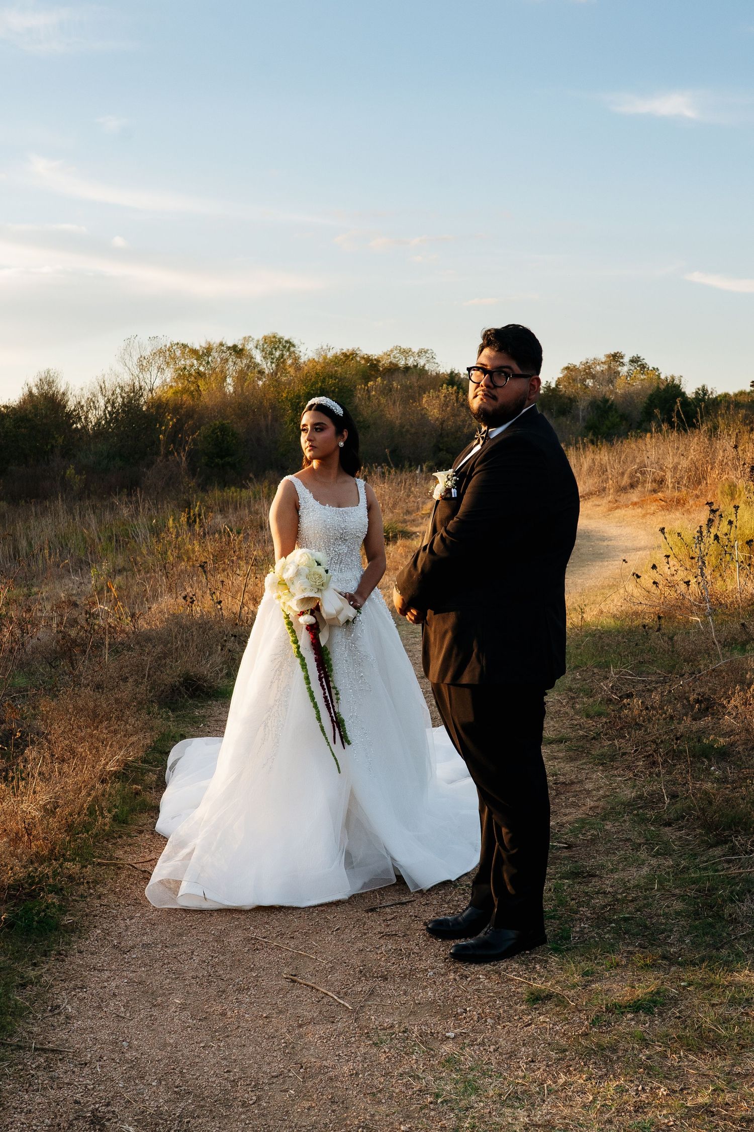 A couple in wedding attire standing on a dirt path during golden hour.