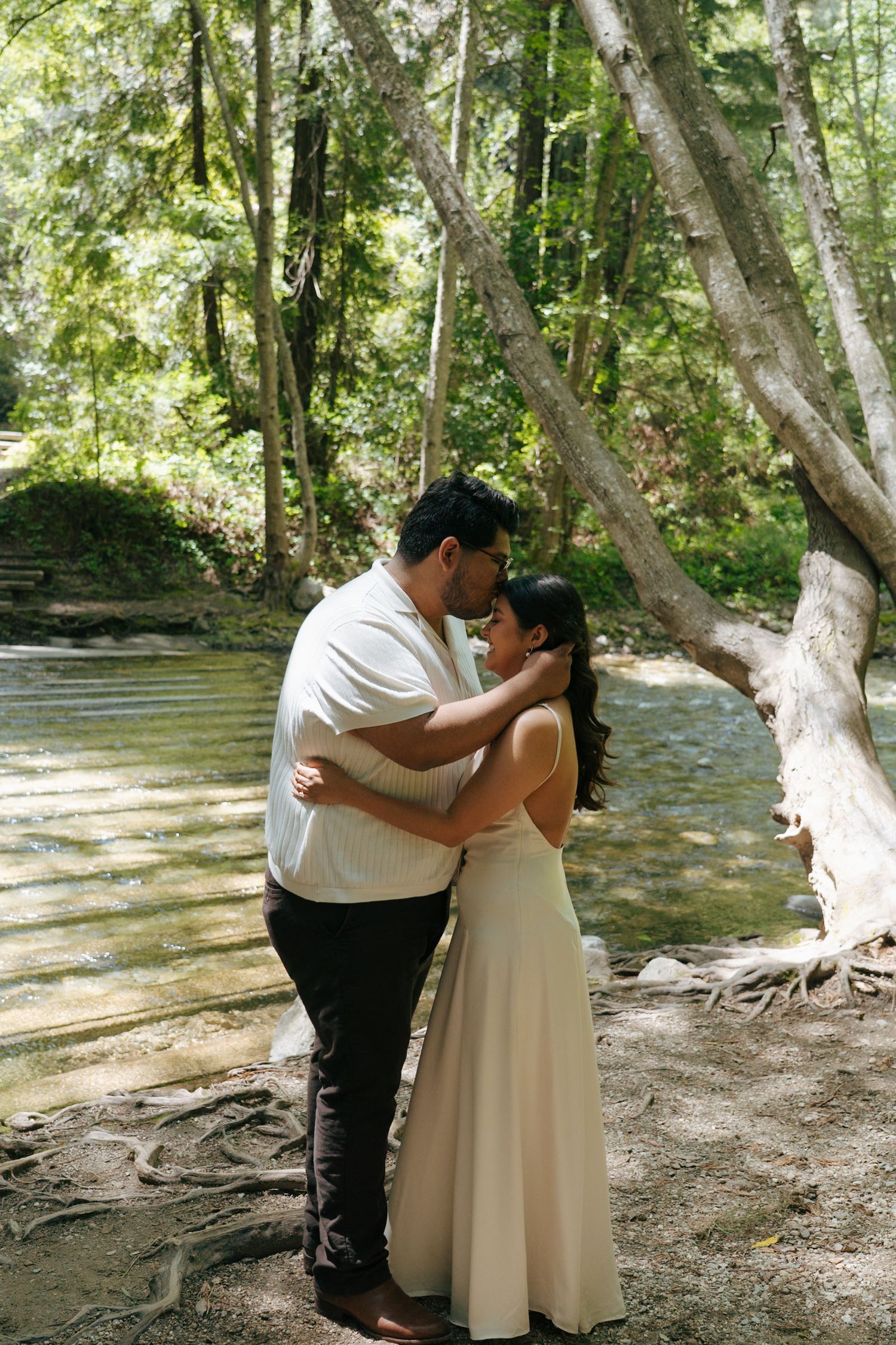 A tender moment between a couple by a stream in a sunlit forest setting.