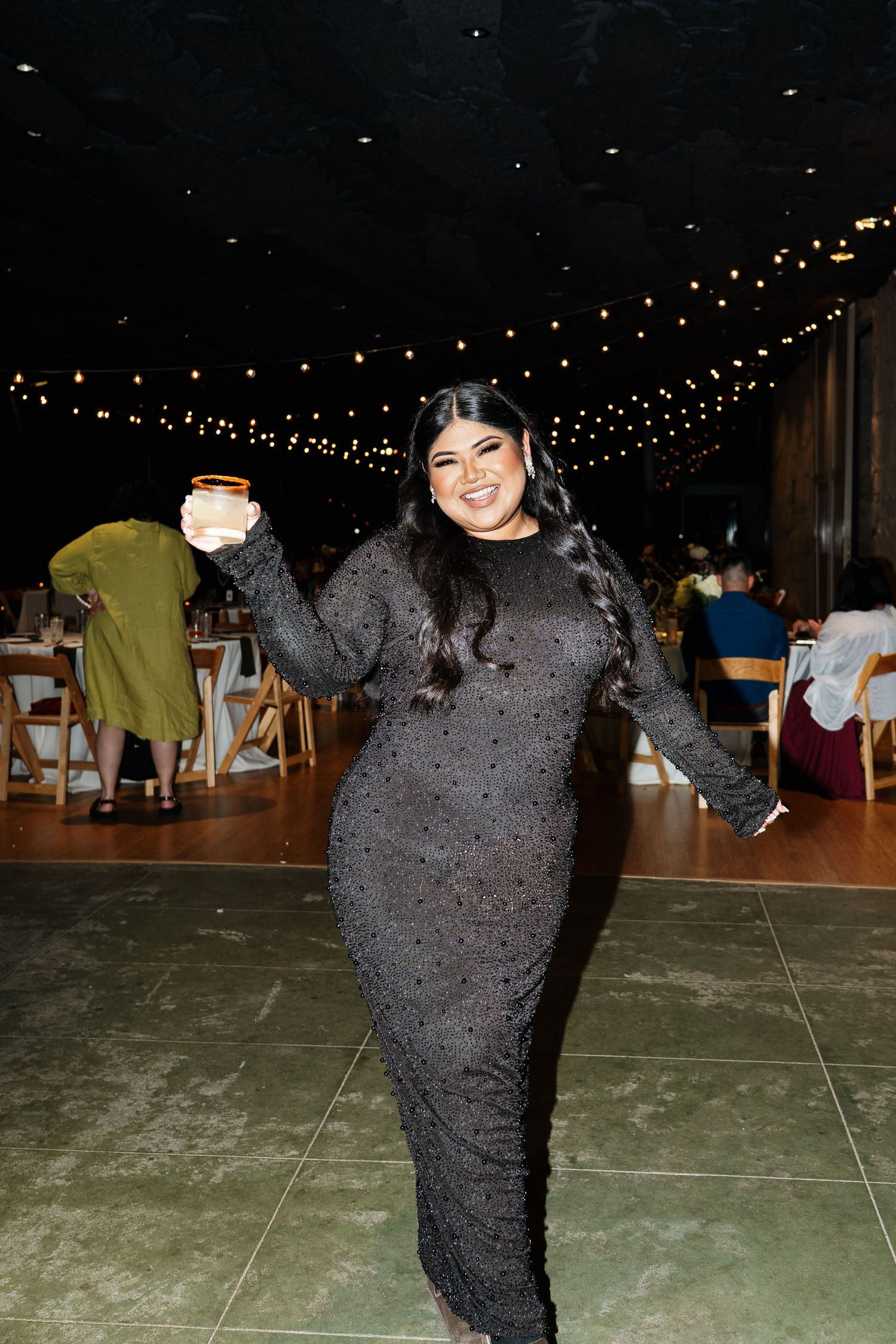 Person in a sparkly dark dress dances under string lights at an evening reception.