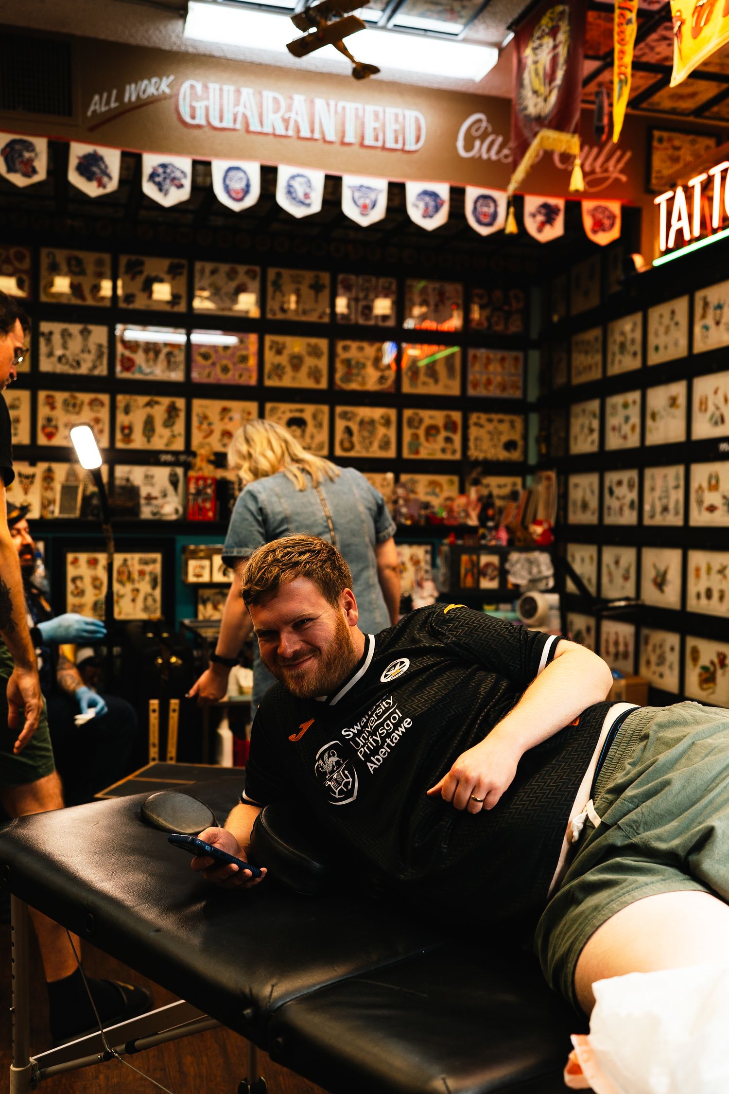 Customer lying on black leather table getting tattooed in traditional tattoo shop setting.
